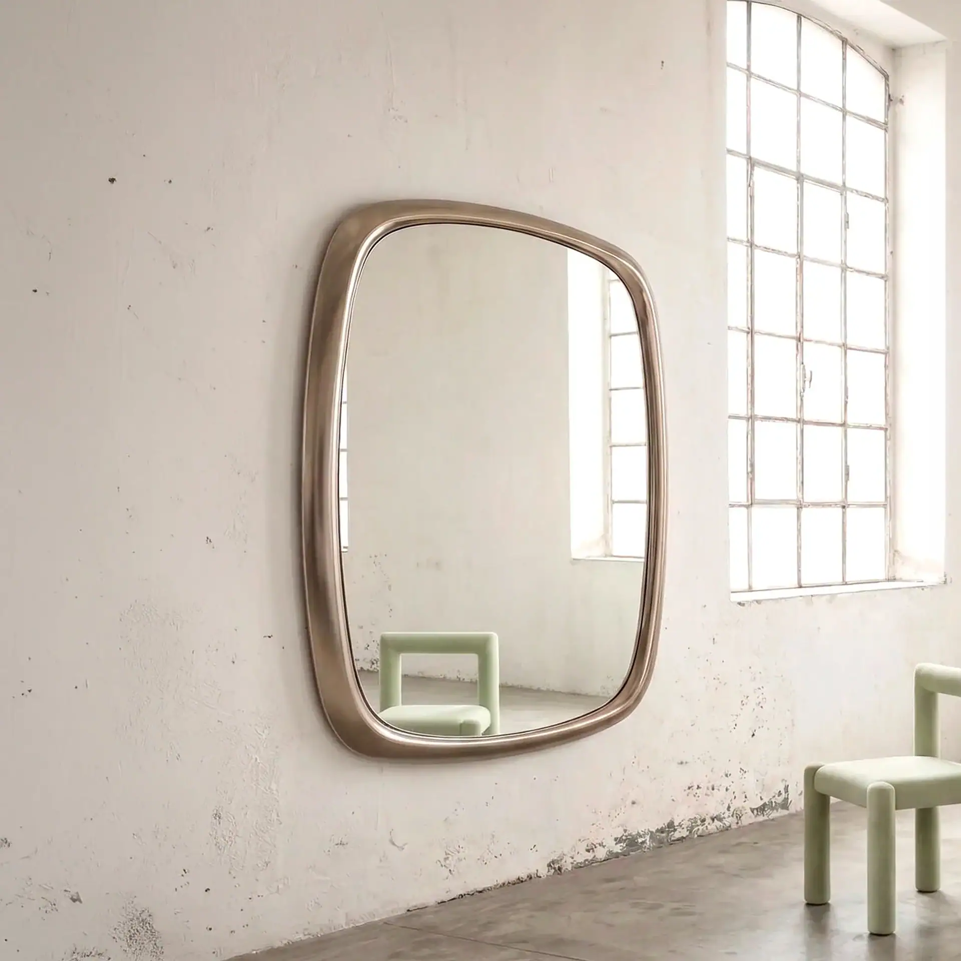 A large, rounded rectangular mirror with a metallic frame on a distressed white wall, reflecting a light green chair and a window. Another similar chair is to the right.