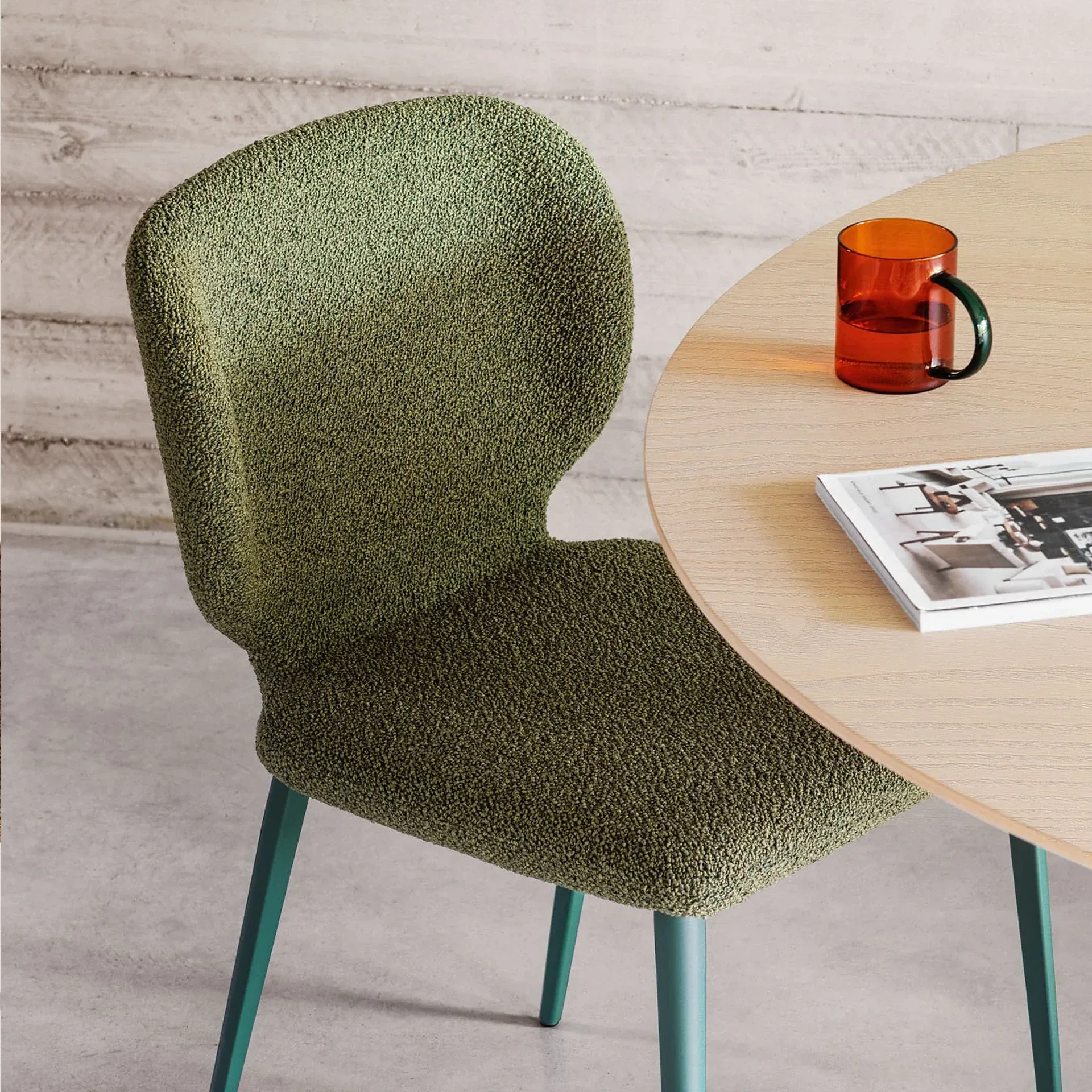 A moss green textured chair next to a light wood round table with an orange glass mug and a magazine.