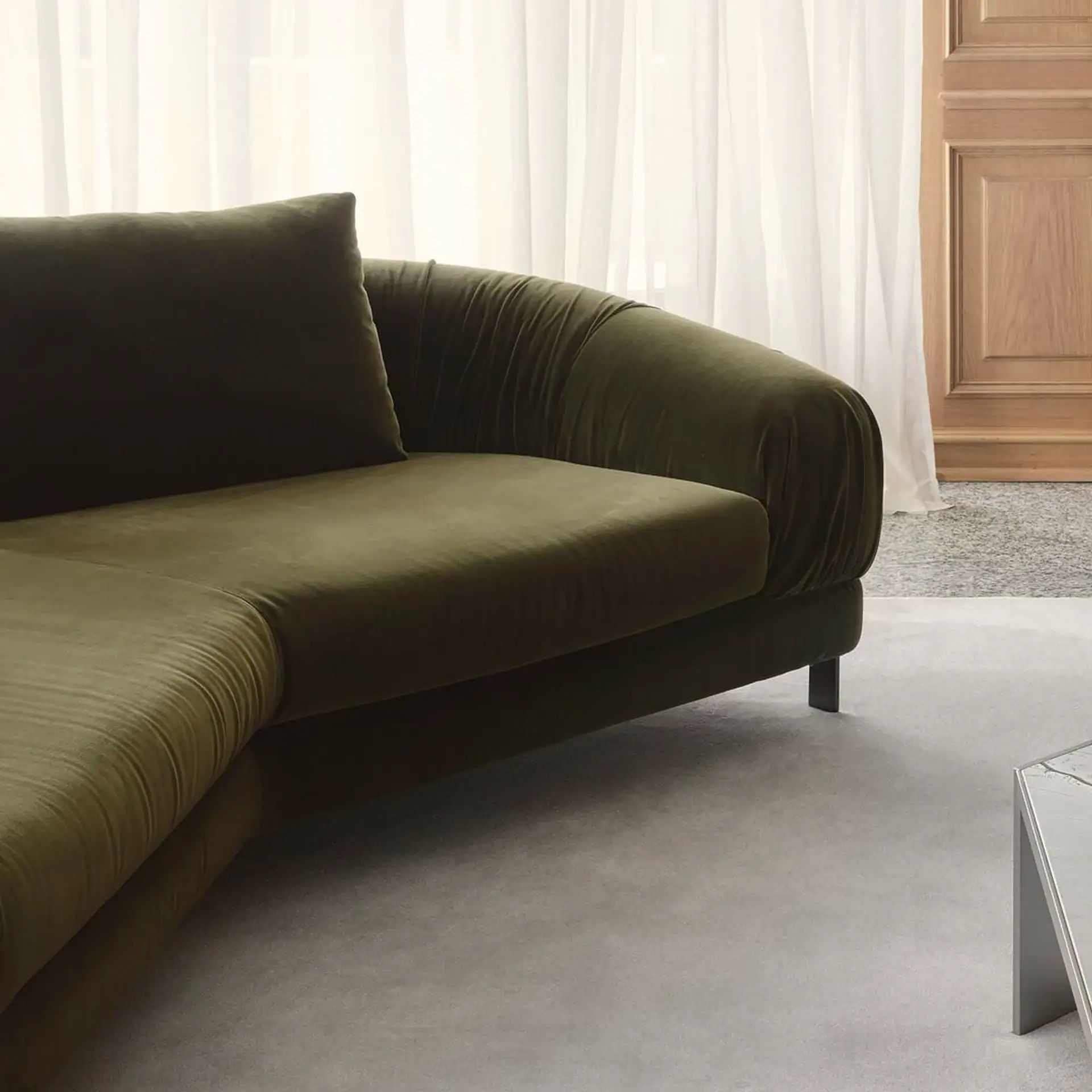 A close-up of an olive green velvet sofa with a curved armrest, set against sheer white curtains and a light gray rug.