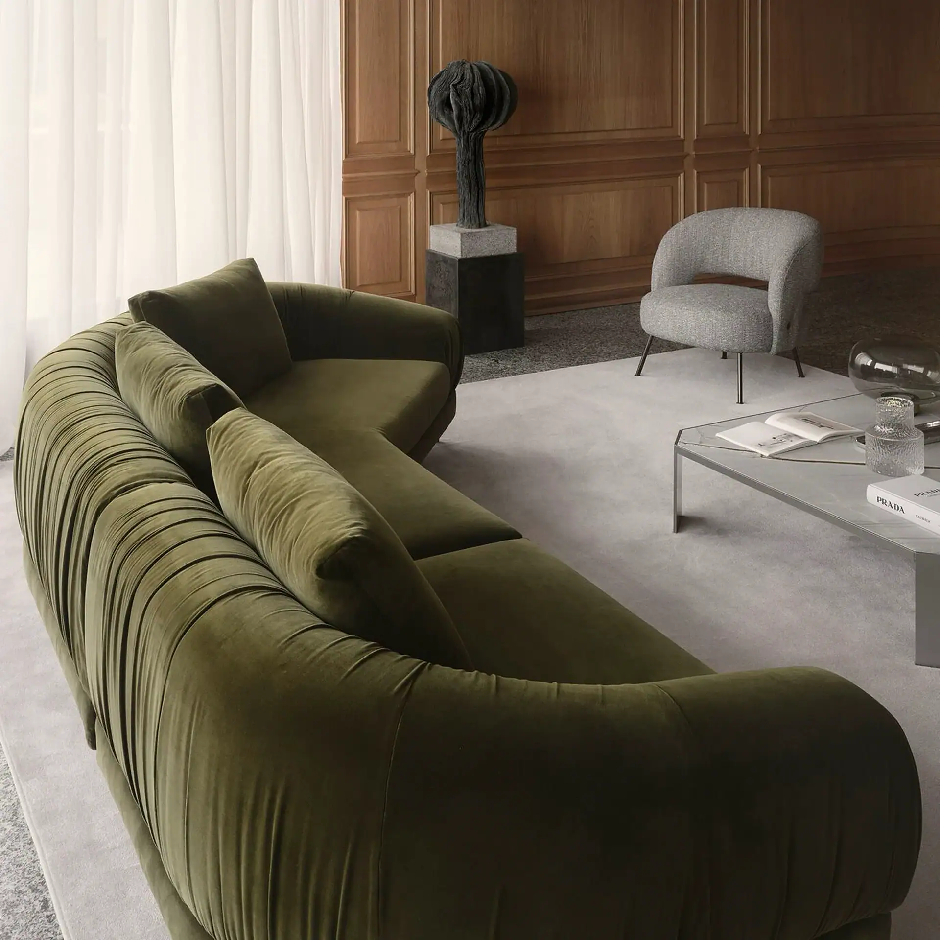 A modern living room with an olive green velvet curved sectional sofa, a grey armchair, a coffee table with books and decor, and a sculptural art piece against a wood-paneled wall.