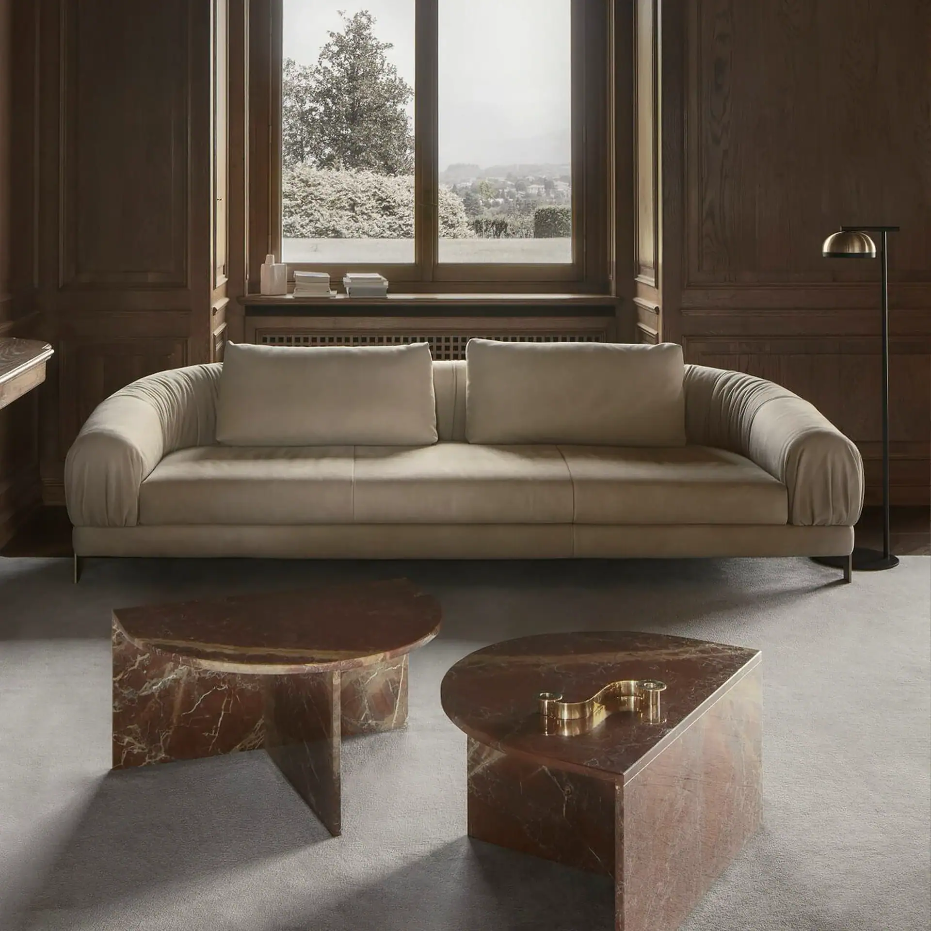 A living room with a beige sofa, two red marble coffee tables, dark wood paneling, and a large window looking out onto a landscape.