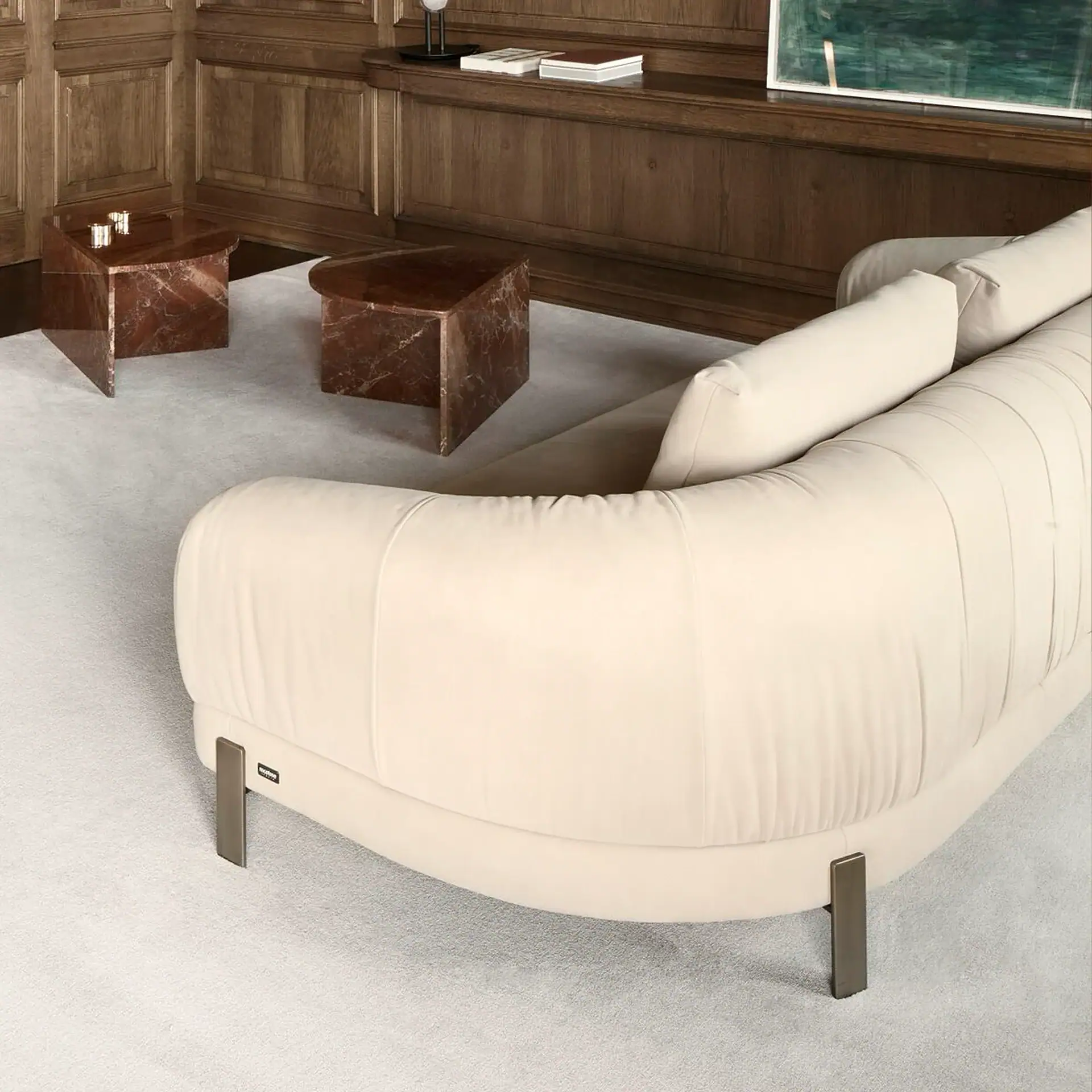 A cream curved sofa with metal legs on a light grey rug, flanked by two angular reddish-brown marble tables. A dark wooden paneled wall with a shelf displays books and a green abstract painting.