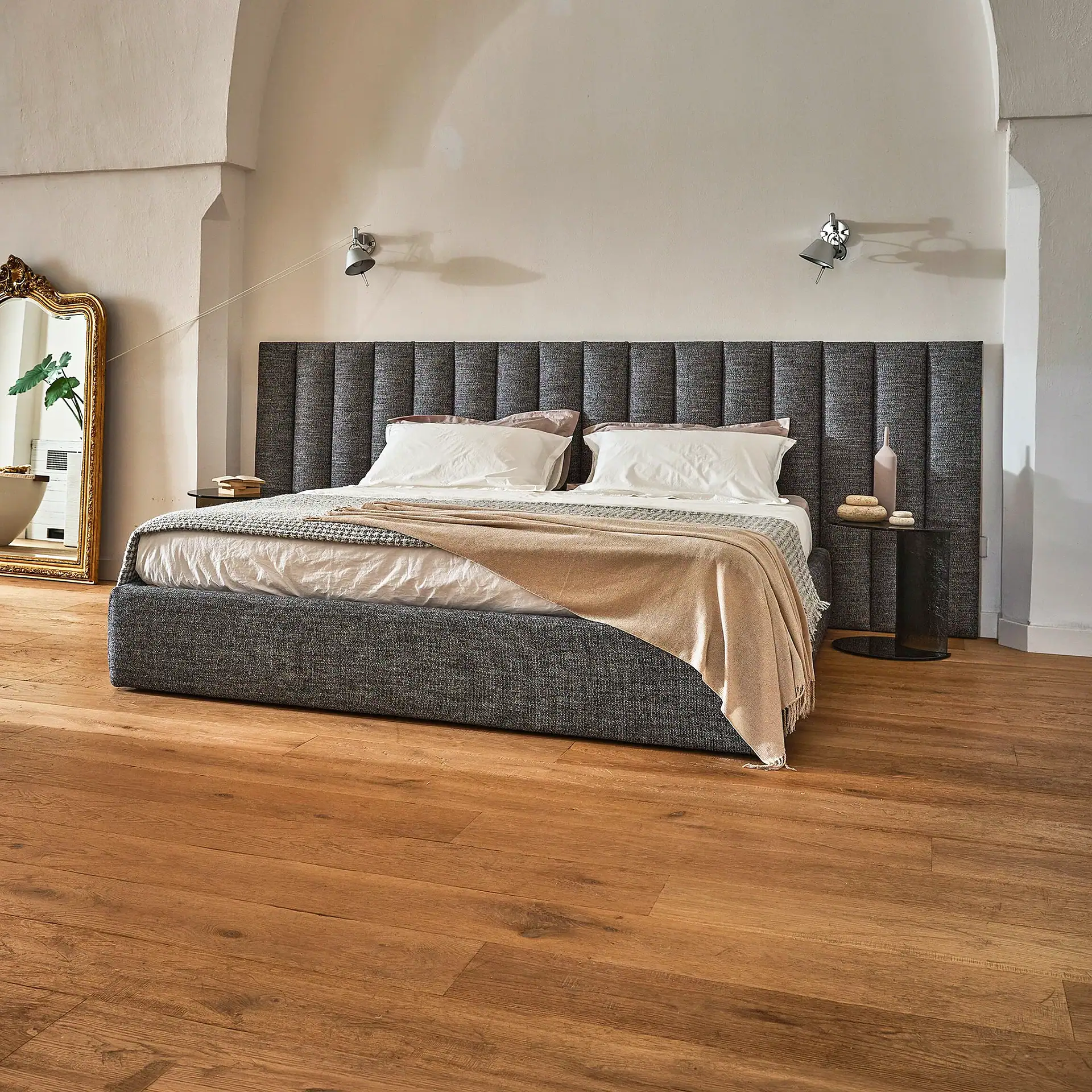 A modern bedroom with a gray upholstered bed, white bedding, wooden floors, and a tall gold-framed mirror.