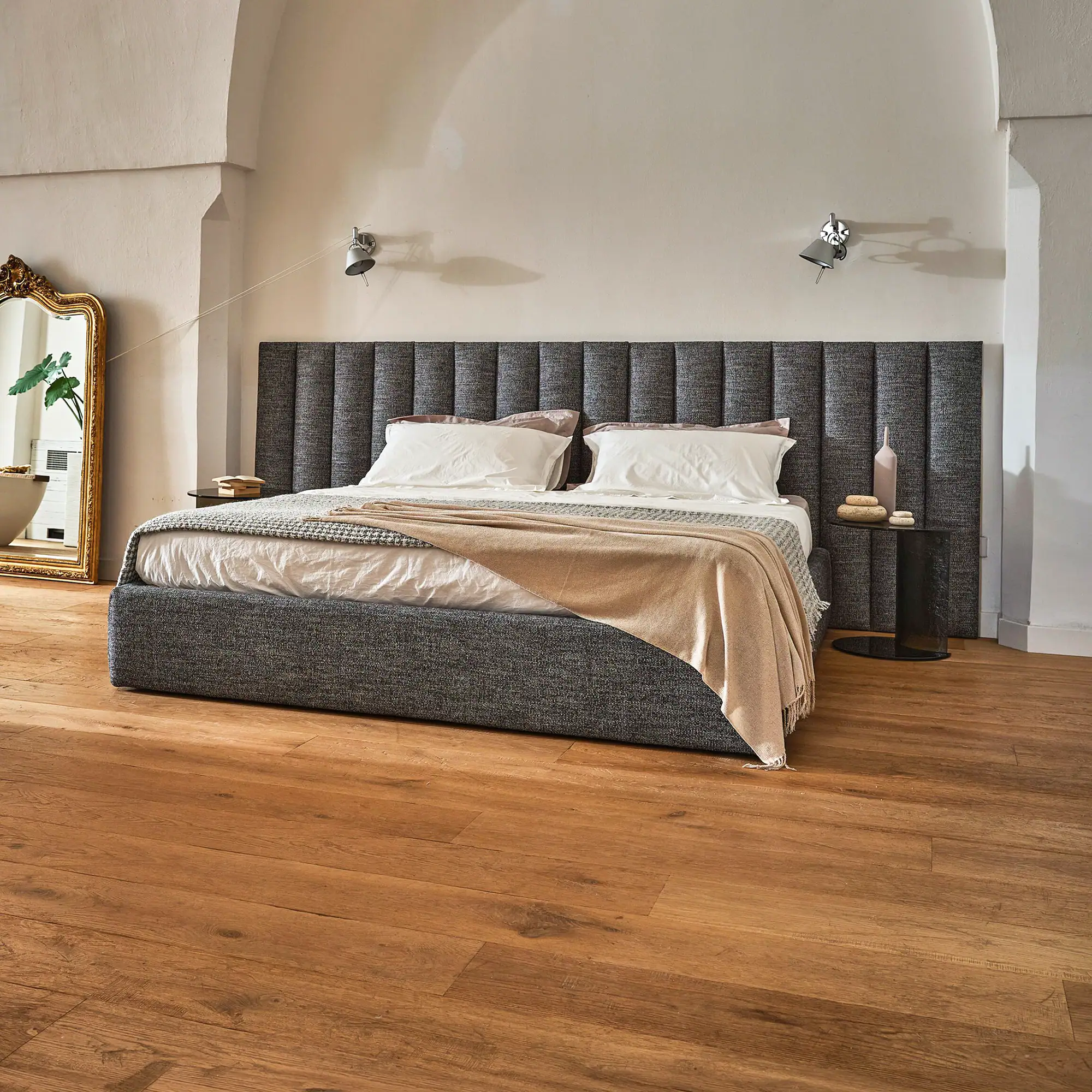 A modern bedroom with a gray upholstered bed, white bedding, wooden floors, and a tall gold-framed mirror.