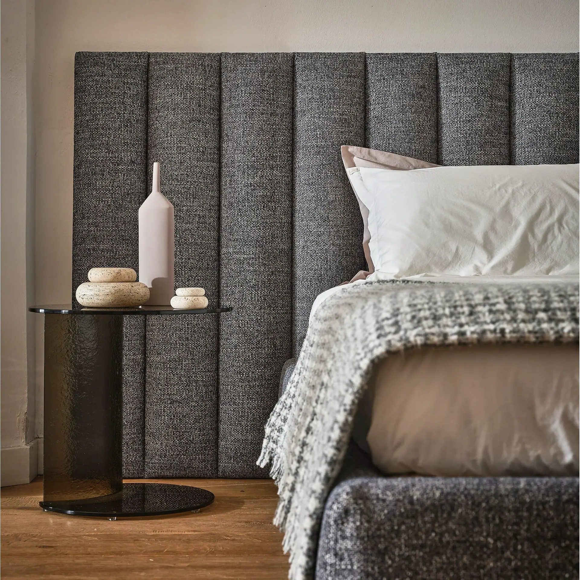 A modern bedroom with a dark grey upholstered headboard, white and pink pillows, and a grey patterned throw blanket. A dark glass bedside table holds a light pink vase and decorative stones.