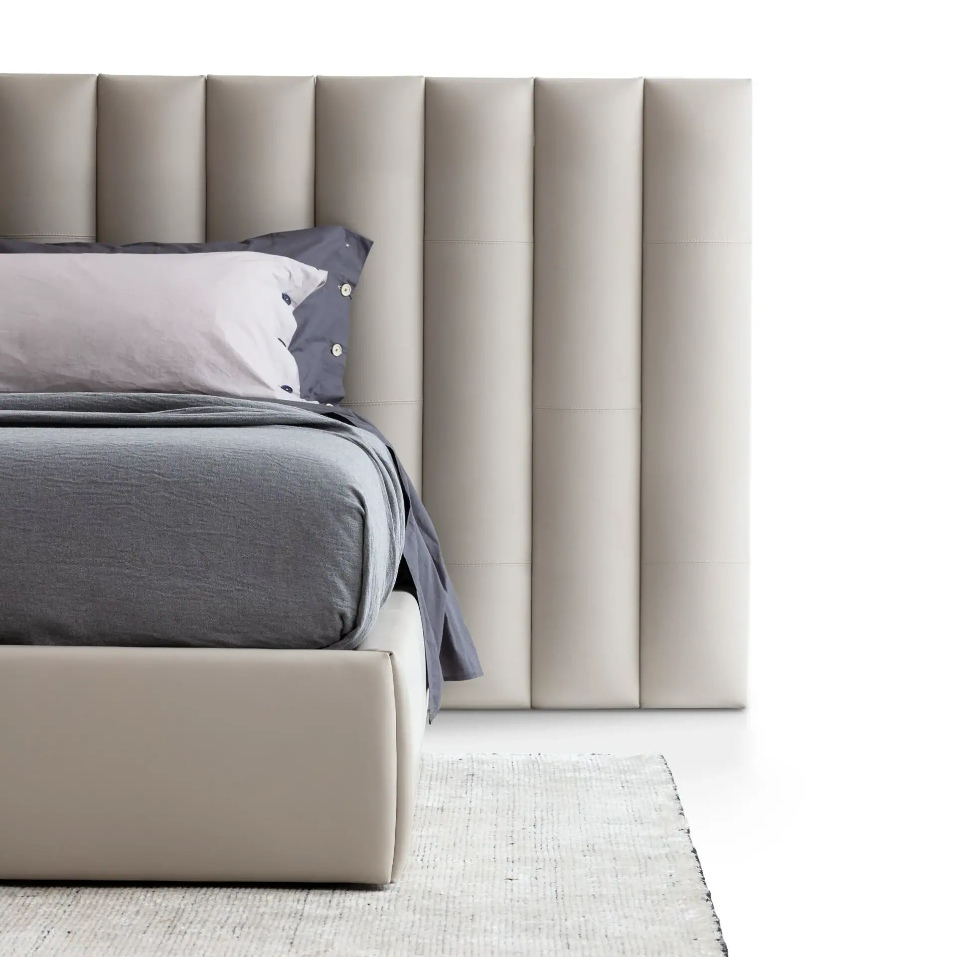 A modern bed with a light beige padded headboard, grey bedding, and a white rug on a white background.