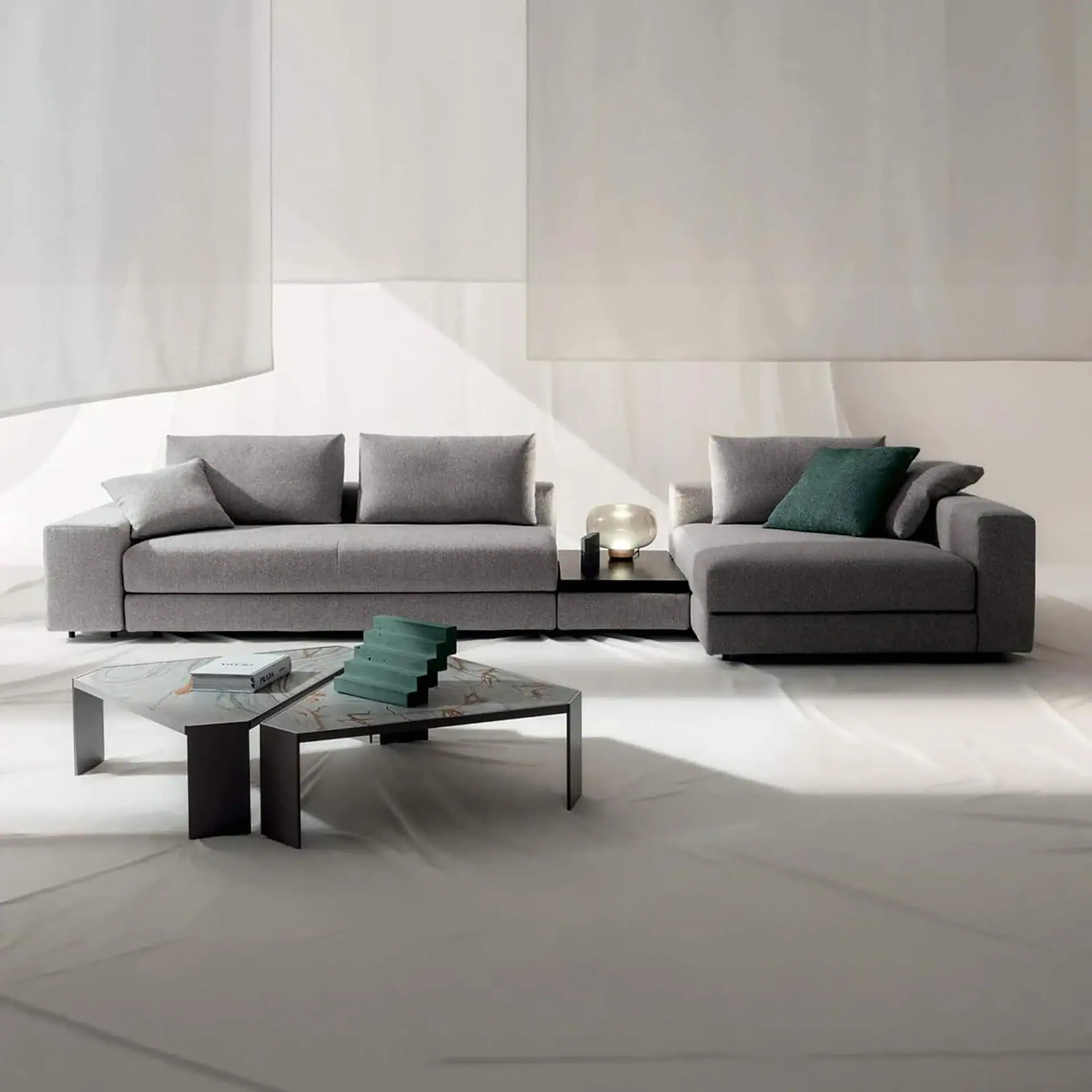 A gray modular sofa with a chaise lounge, connected by a dark table with a glowing lamp. Two hexagonal coffee tables with a book and green decor are in front. The setting is a minimalist studio with a light background.