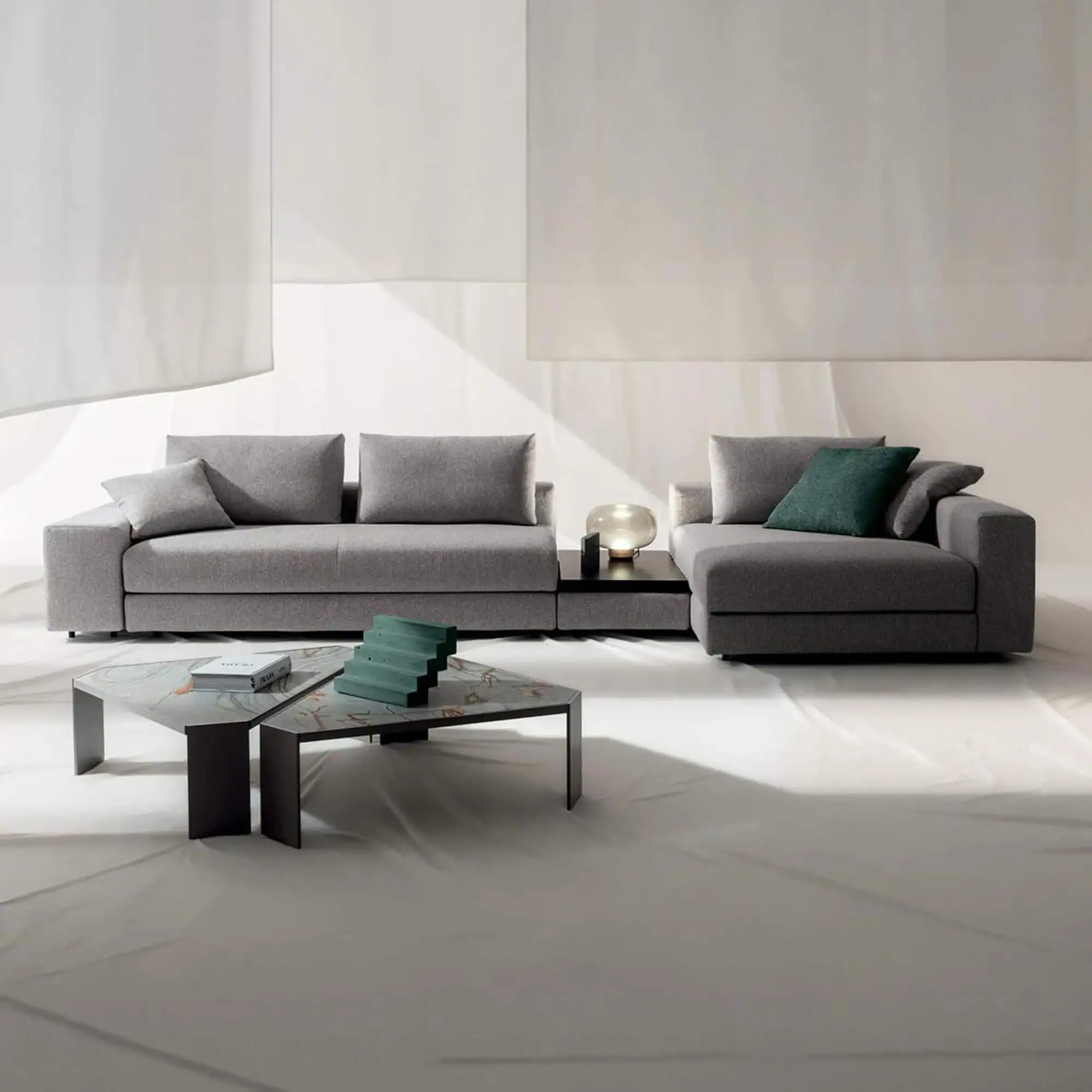 A gray modular sofa with a chaise lounge, connected by a dark table with a glowing lamp. Two hexagonal coffee tables with a book and green decor are in front. The setting is a minimalist studio with a light background.
