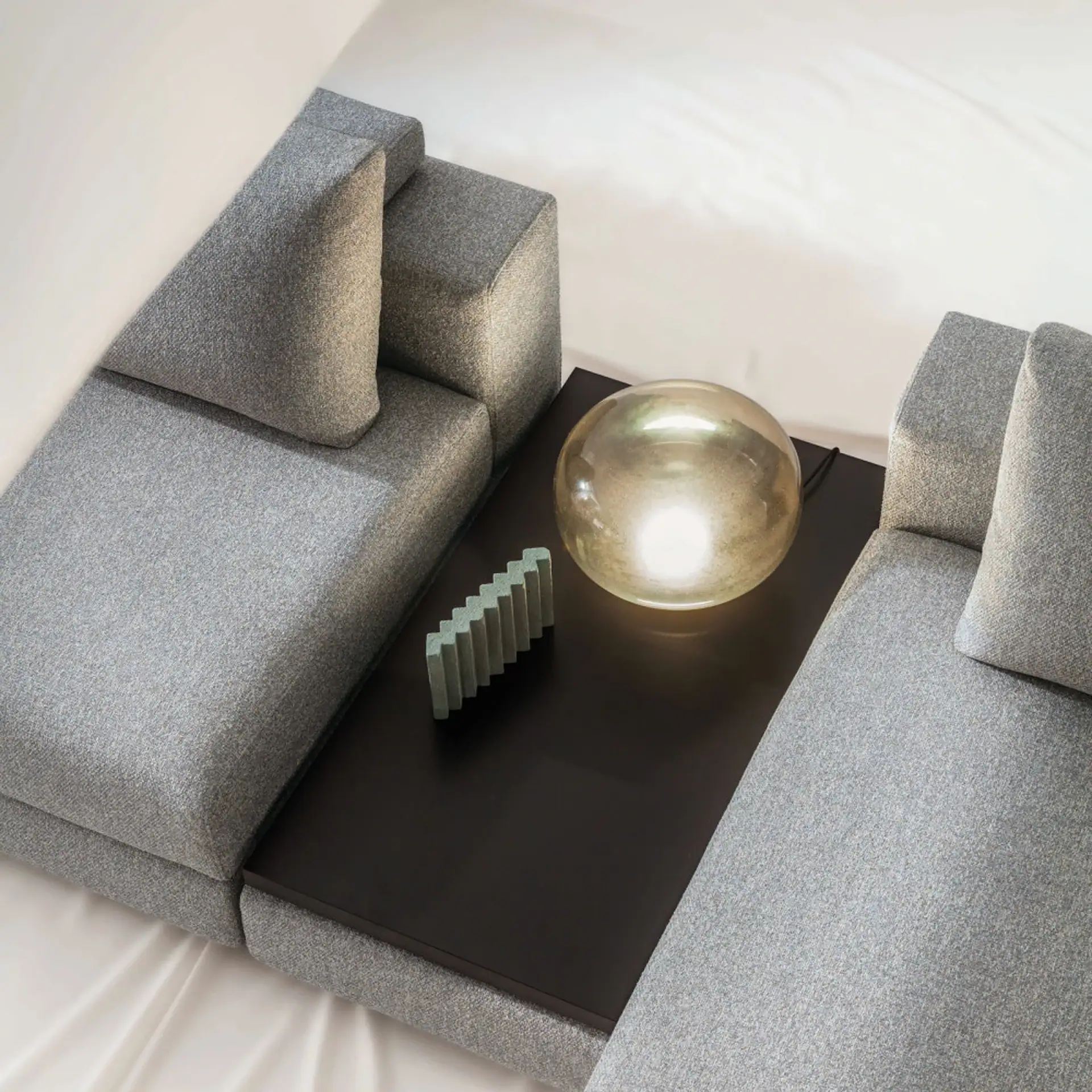 Top-down view of a grey modular sofa with an integrated dark table, a glowing spherical amber lamp, and a light green stepped decorative object.