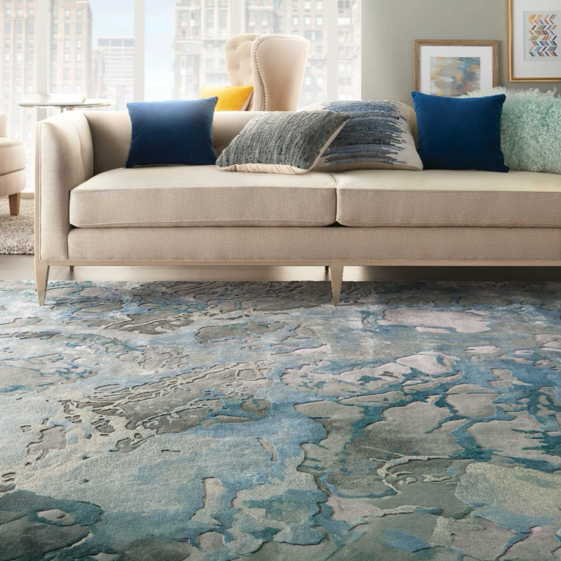 A modern living room with a blue and grey abstract rug, a light beige sofa with blue and grey pillows, and two armchairs with a yellow pillow, set against a city window view.