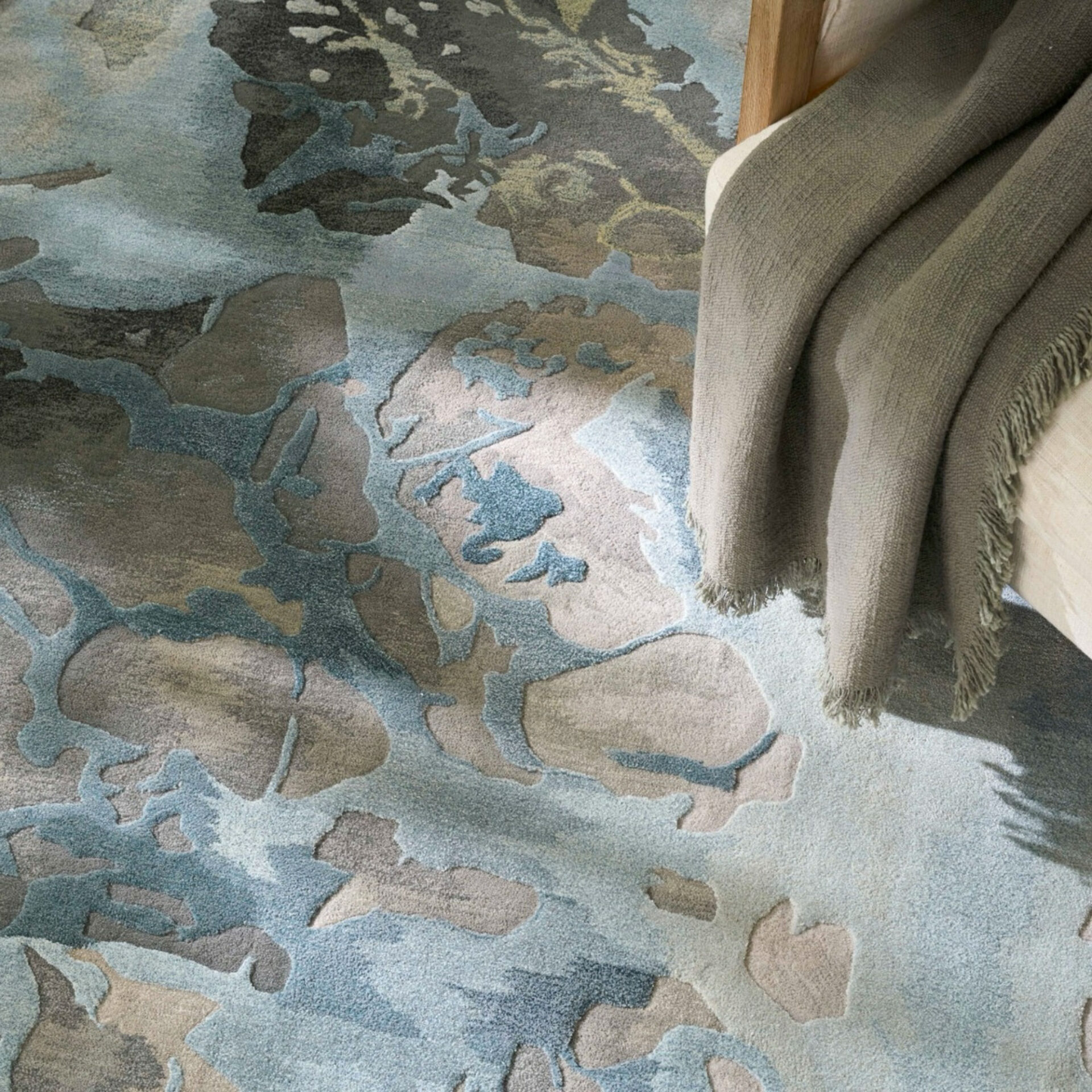 An abstract patterned rug in shades of blue, grey, and beige with organic shapes, seen from above. A light grey fringed blanket is draped over a chair in the upper right.
