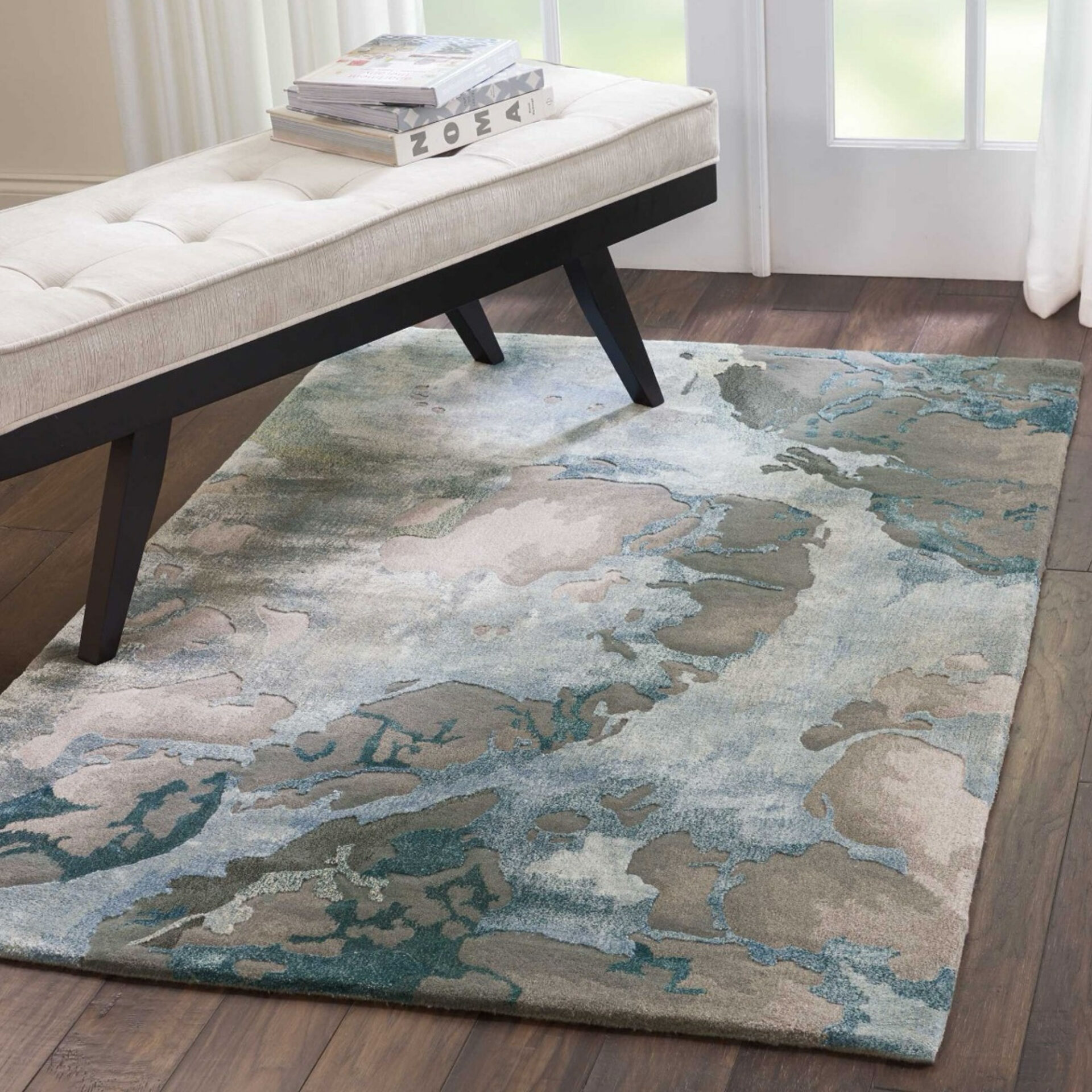 A rectangular area rug with an abstract pattern in blue, teal, gray, and beige on dark wood flooring. A cream tufted bench with dark legs sits on the rug, holding a stack of books. A window with sheer curtains is in the background.