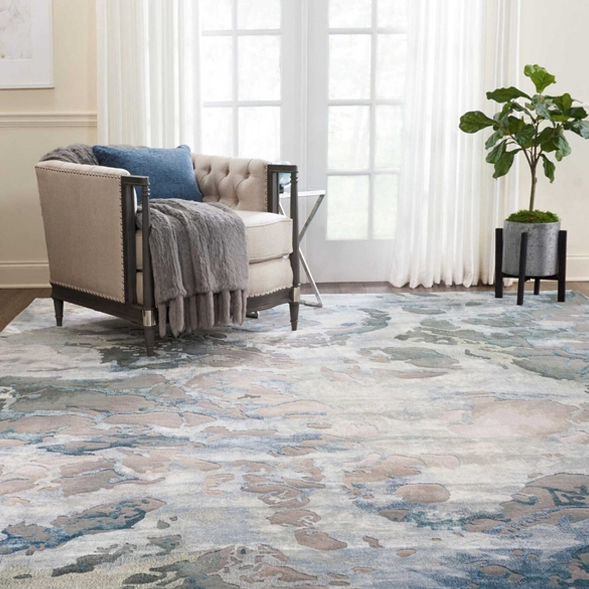 A cozy living room with a light beige armchair, a blue and grey abstract patterned rug, a large window with sheer white curtains, and a potted Fiddle Leaf Fig plant.