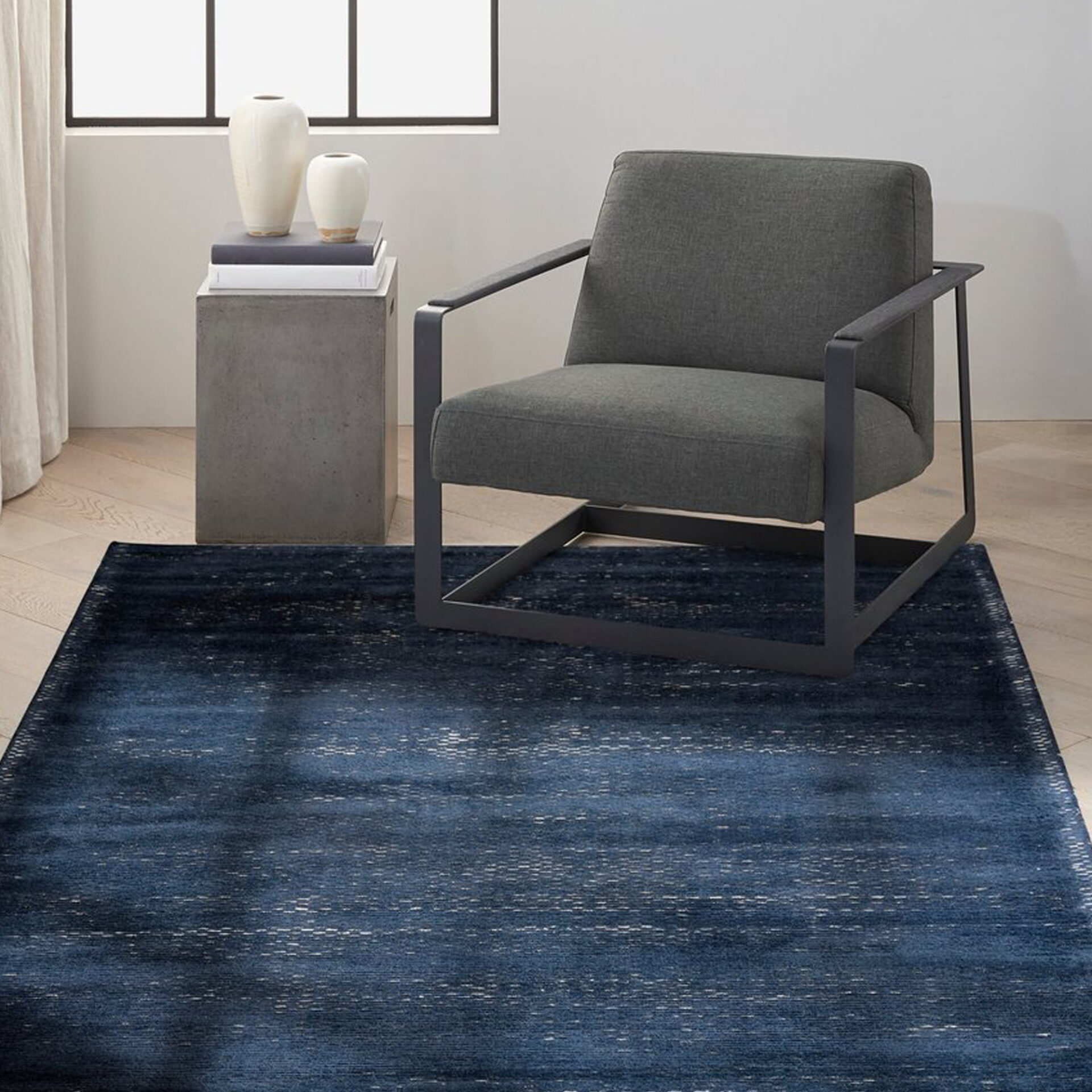 A modern living space featuring a deep indigo blue area rug with abstract patterns. A dark grey armchair and a concrete side table with two white vases sit on the rug, against a light wall with a window.