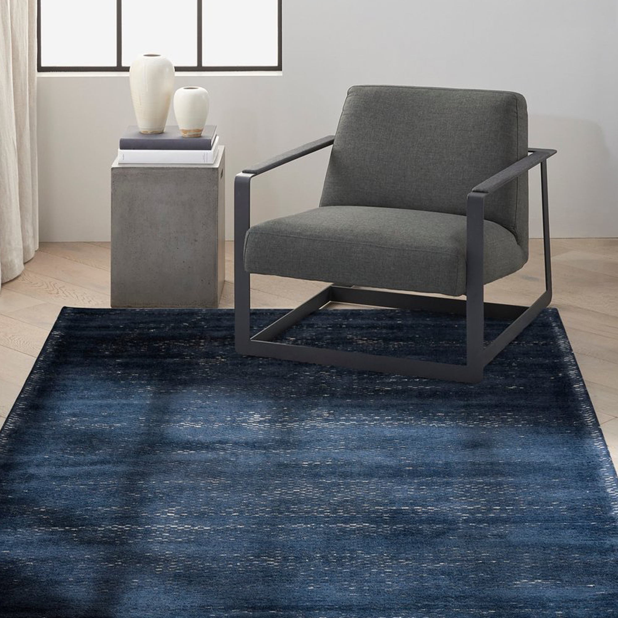 A modern living space featuring a deep indigo blue area rug with abstract patterns. A dark grey armchair and a concrete side table with two white vases sit on the rug, against a light wall with a window.