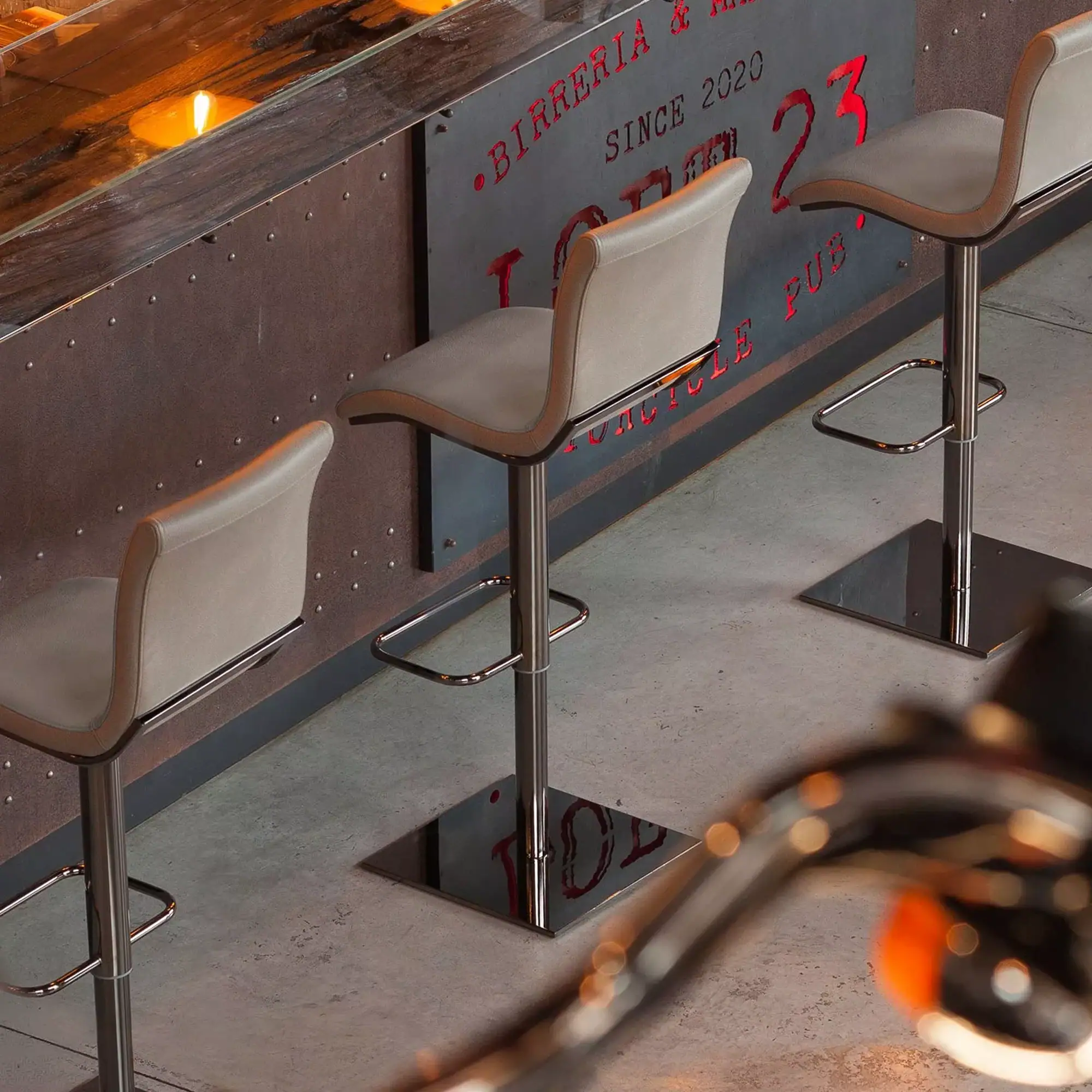 Three beige bar stools with chrome bases in front of a dark, riveted metal bar with a red backlit sign. The floor is concrete.