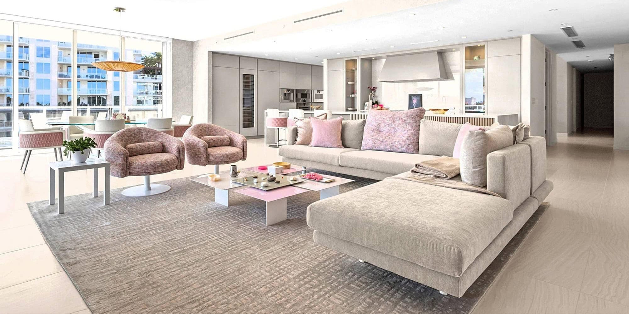 A modern open-concept living room, dining area, and kitchen. The space features light beige and pink furniture, including an L-shaped sofa, two armchairs, and a glass dining table. Large windows overlook apartment buildings, and the kitchen has sleek, light gray cabinetry.