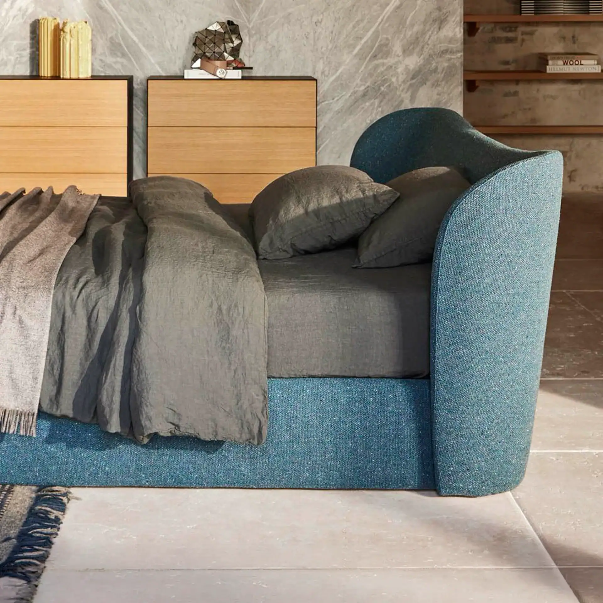 A modern bedroom with a teal blue upholstered bed, gray linen bedding, wooden nightstands, and a marble-patterned wall.