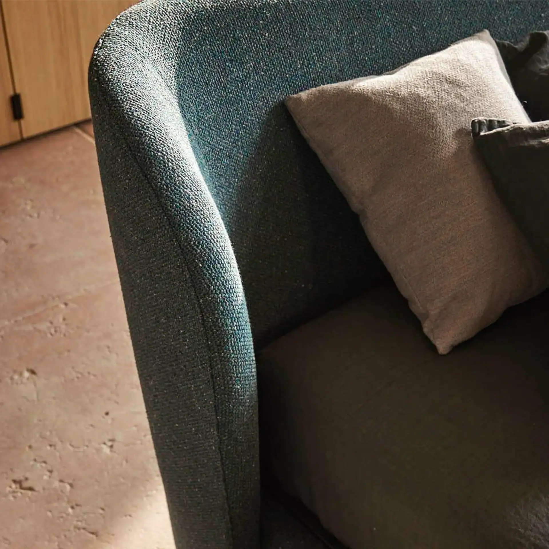 Close-up of a blue-green textured sofa armrest with a light brown throw pillow and a darker cushion, next to a wooden panel and textured floor.