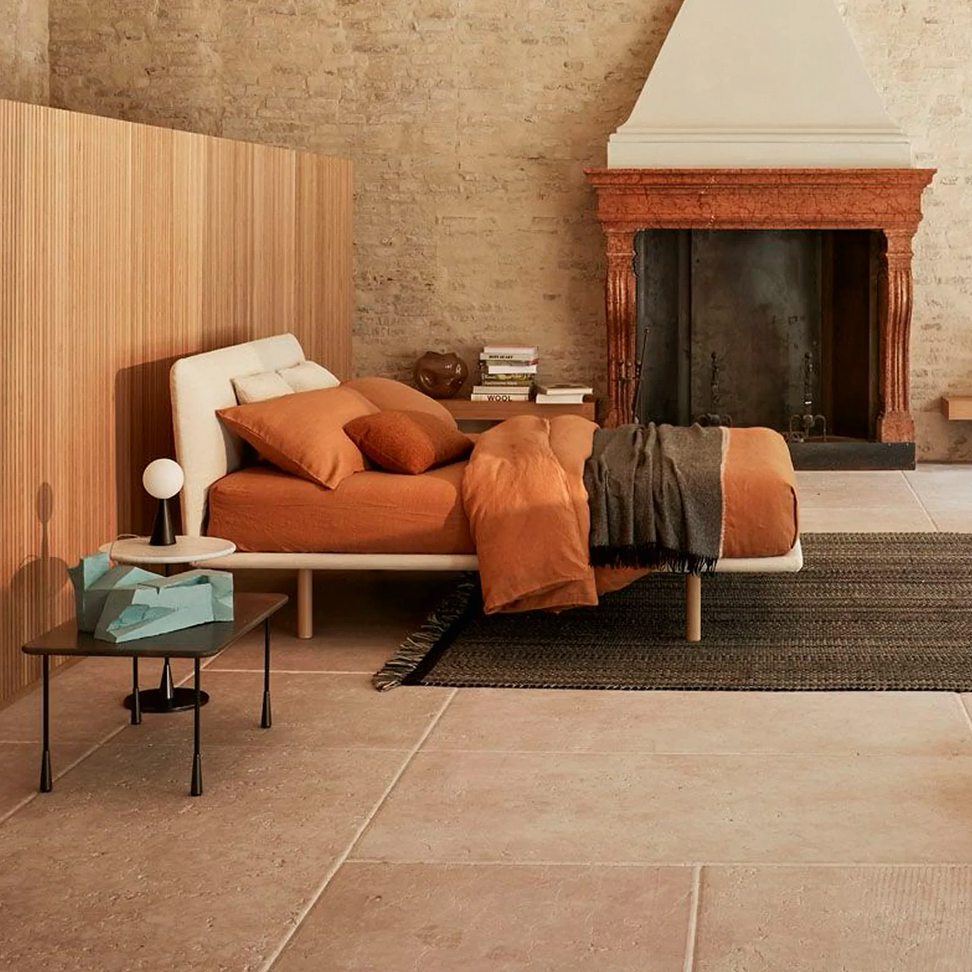 A modern bedroom featuring a bed with orange bedding, a fluted wooden wall panel, a rustic stone fireplace, and a stone-tiled floor with a dark rug.