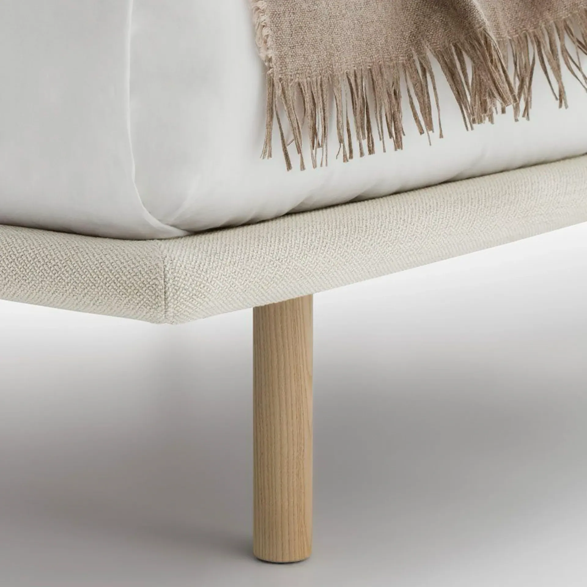 Close-up of a bed showing a light beige upholstered frame, white mattress, fringed brown blanket, and a cylindrical light wood leg.