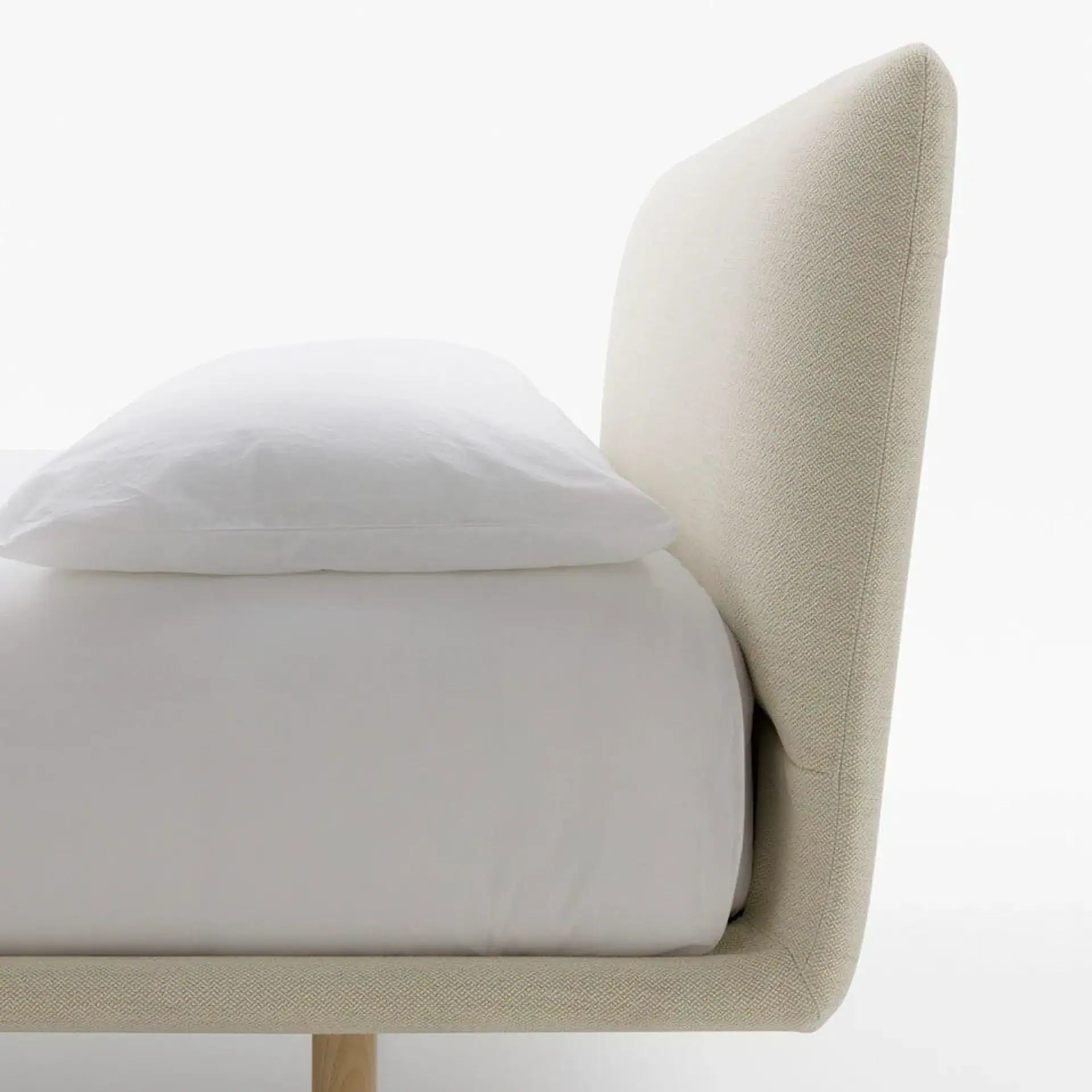 Side view of a bed with a curved, light beige upholstered headboard and frame, a white fitted sheet, and a white pillow.