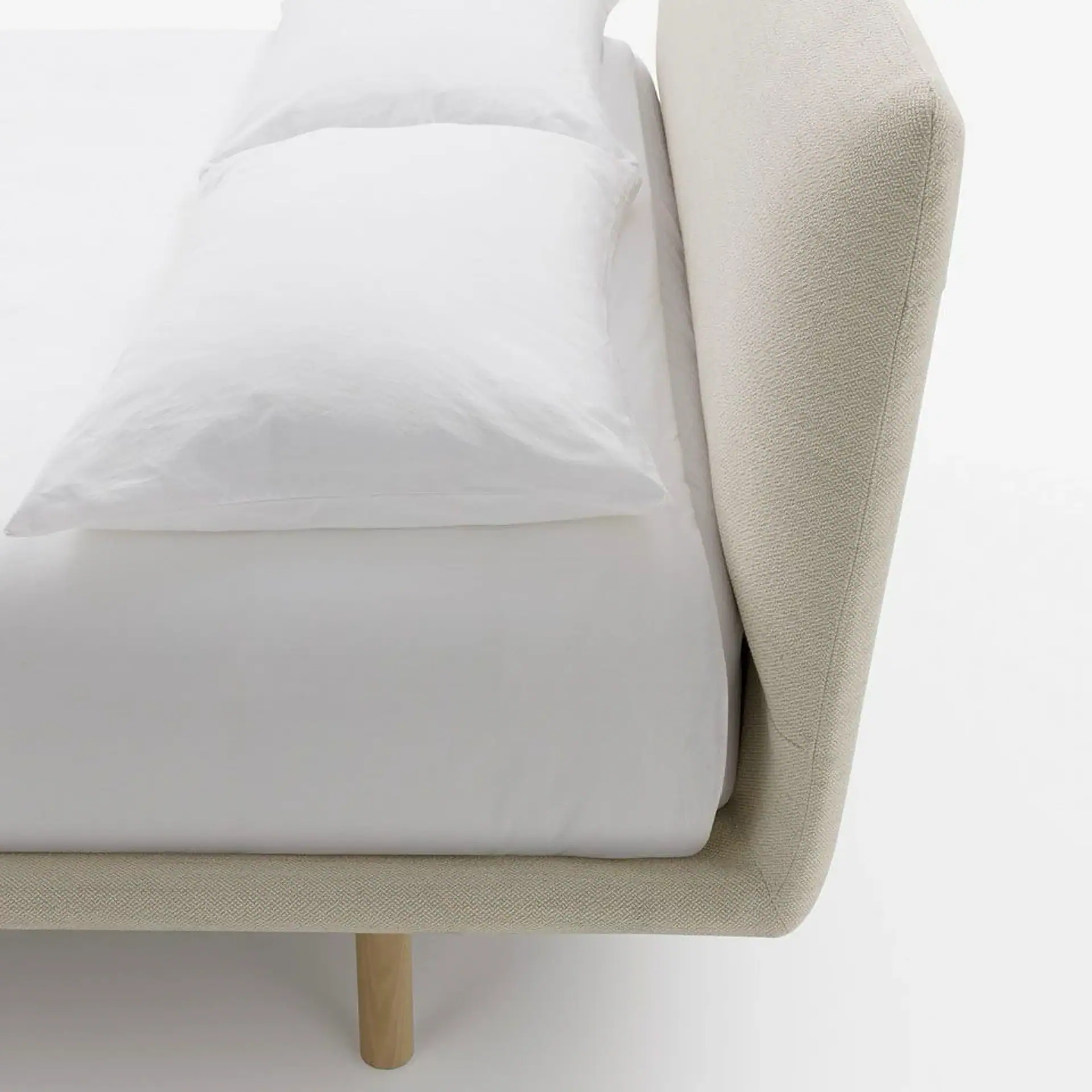 Close-up of a bed with a beige fabric headboard, white sheets, and two white pillows, with a wooden leg visible.
