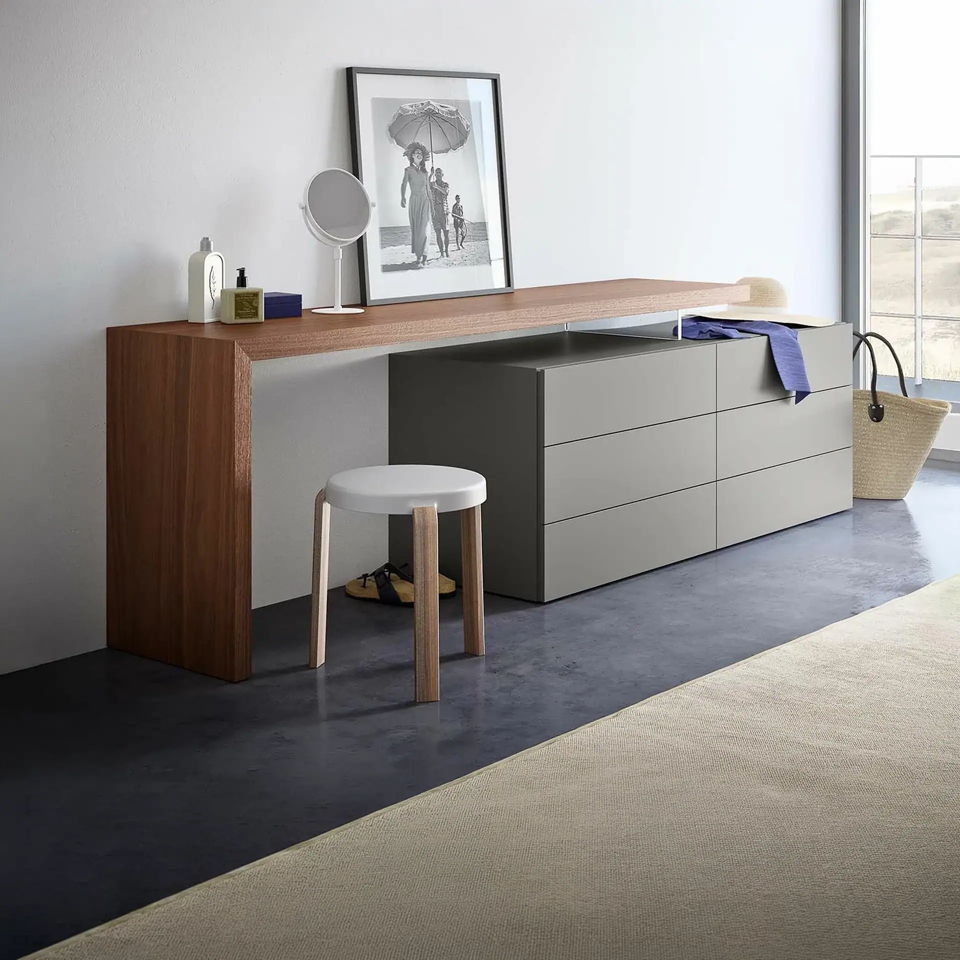 A modern dressing table setup with a wooden console, grey three-drawer dresser, white stool, vanity mirror, framed beach photo, and a window view.