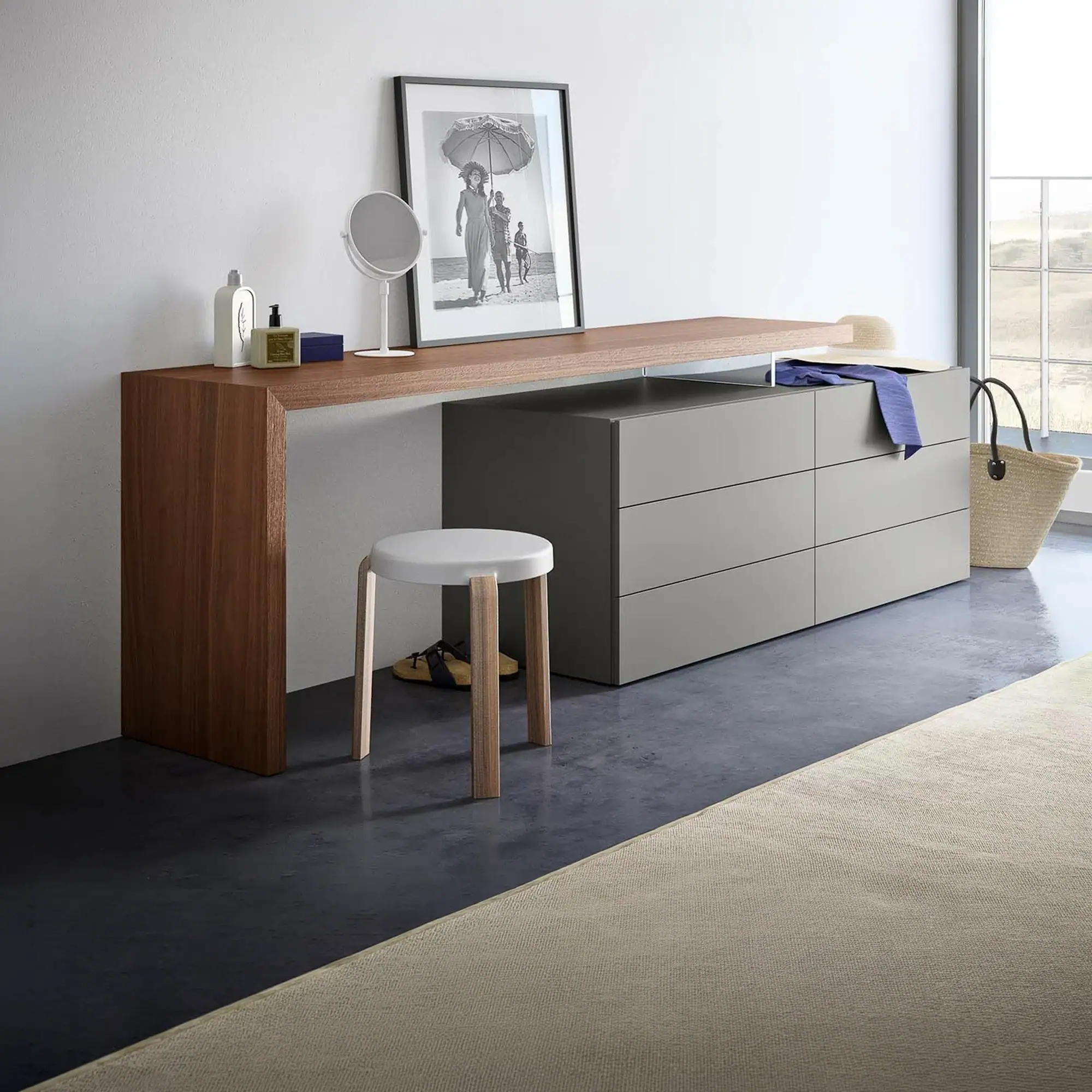 A modern dressing table setup with a wooden console, grey three-drawer dresser, white stool, vanity mirror, framed beach photo, and a window view.