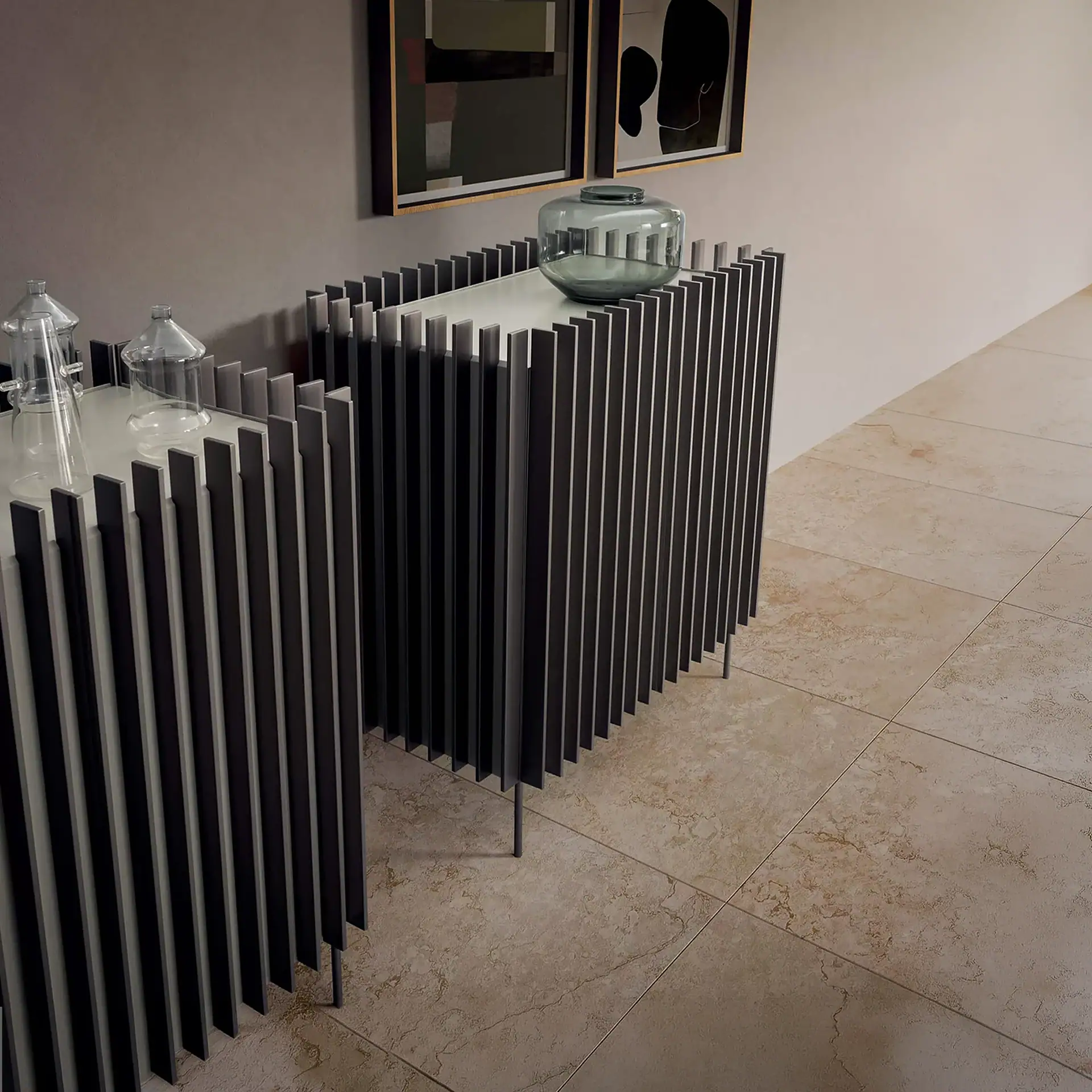 Two modern console tables with slatted dark grey fronts and light tops. One has a clear glass cloche, the other a dark grey glass vase. Beige tiled floor and grey wall with two framed abstract artworks.