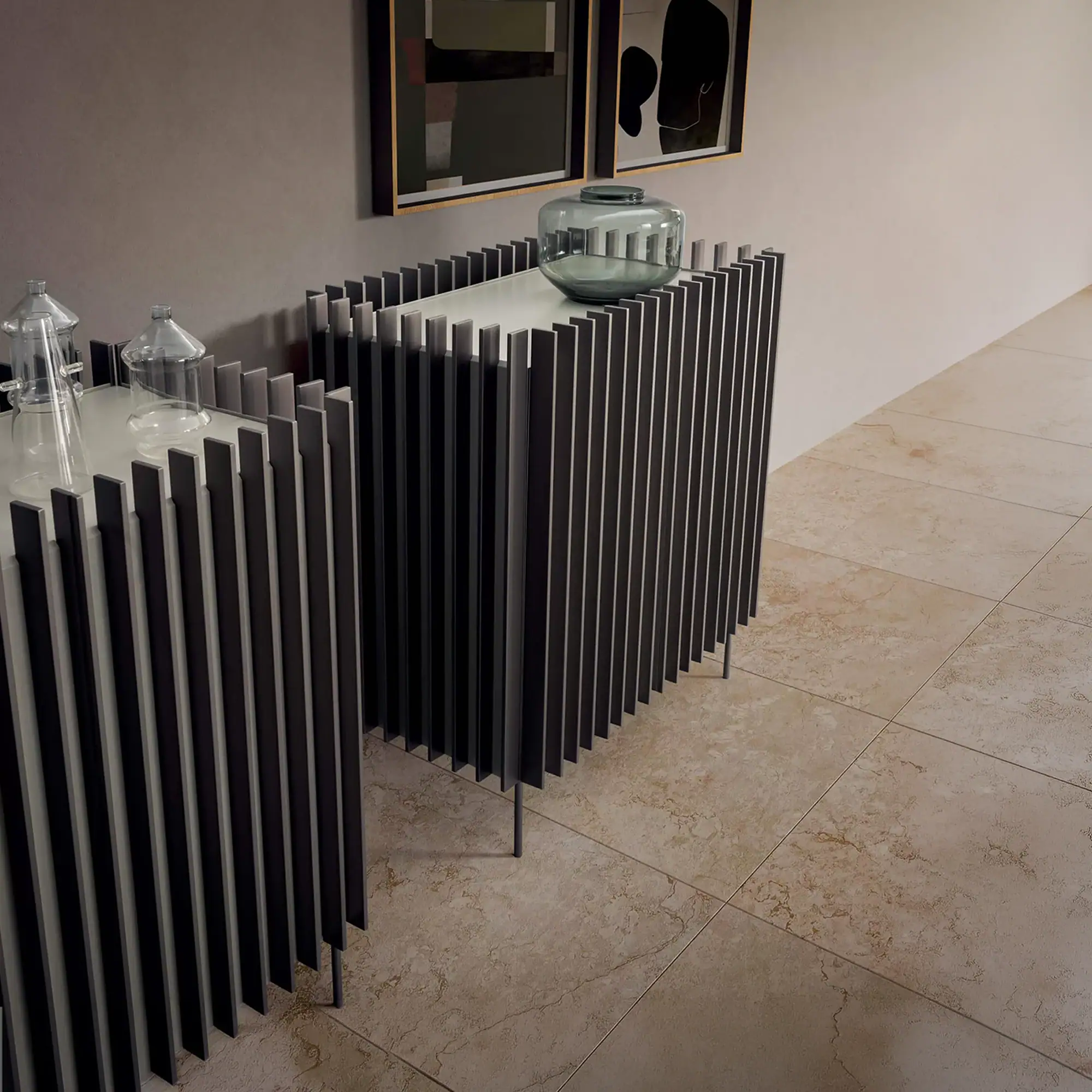 Two modern console tables with slatted dark grey fronts and light tops. One has a clear glass cloche, the other a dark grey glass vase. Beige tiled floor and grey wall with two framed abstract artworks.