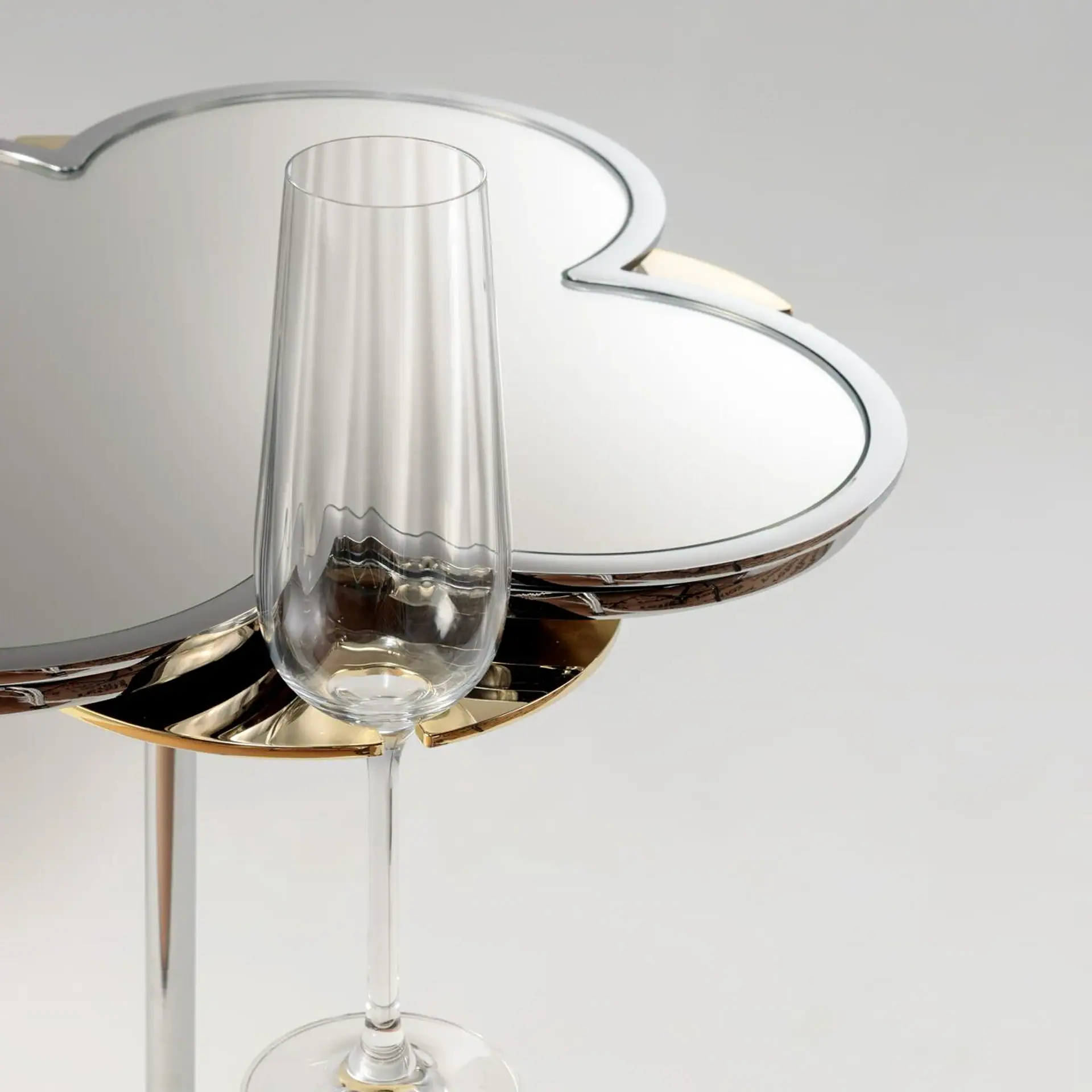 A clear champagne flute stands in front of a quatrefoil-shaped mirrored table with silver and gold accents, against a light gray background.