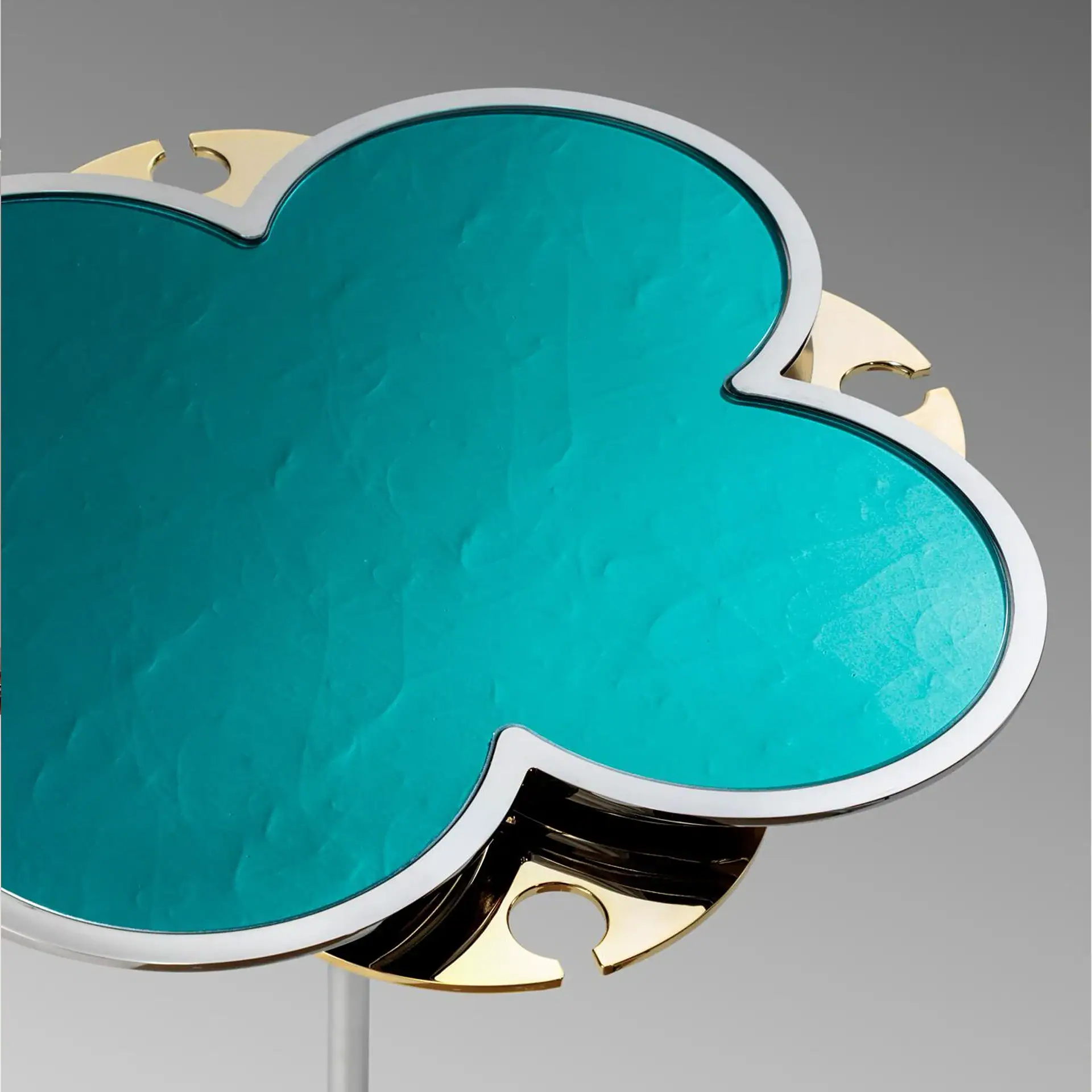 Close-up of a decorative object shaped like a four-leaf clover, with a textured teal surface, silver frame, and gold metallic backing, against a gray background.