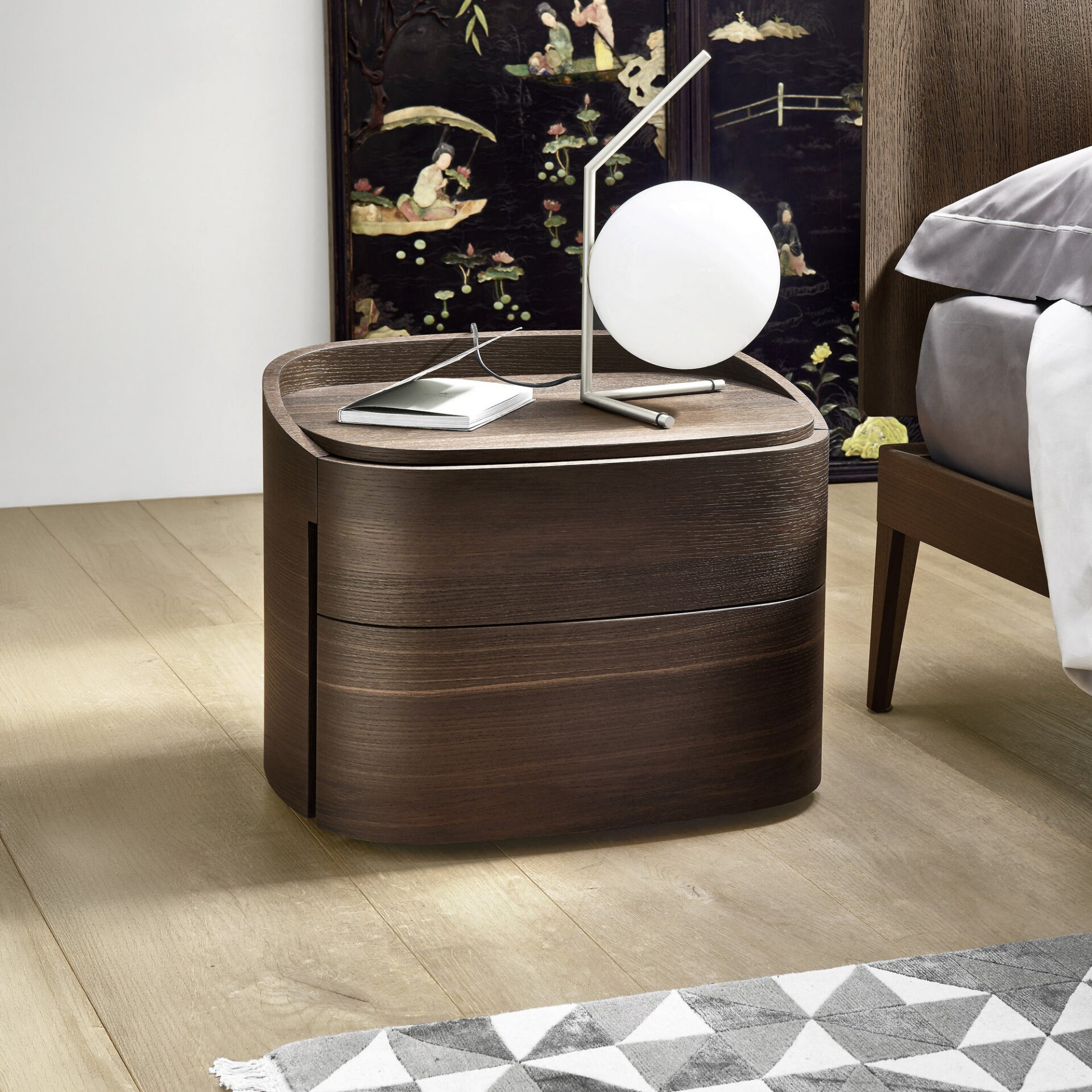 A modern dark wood nightstand with a white spherical lamp and a book, next to a bed and an ornate black screen.