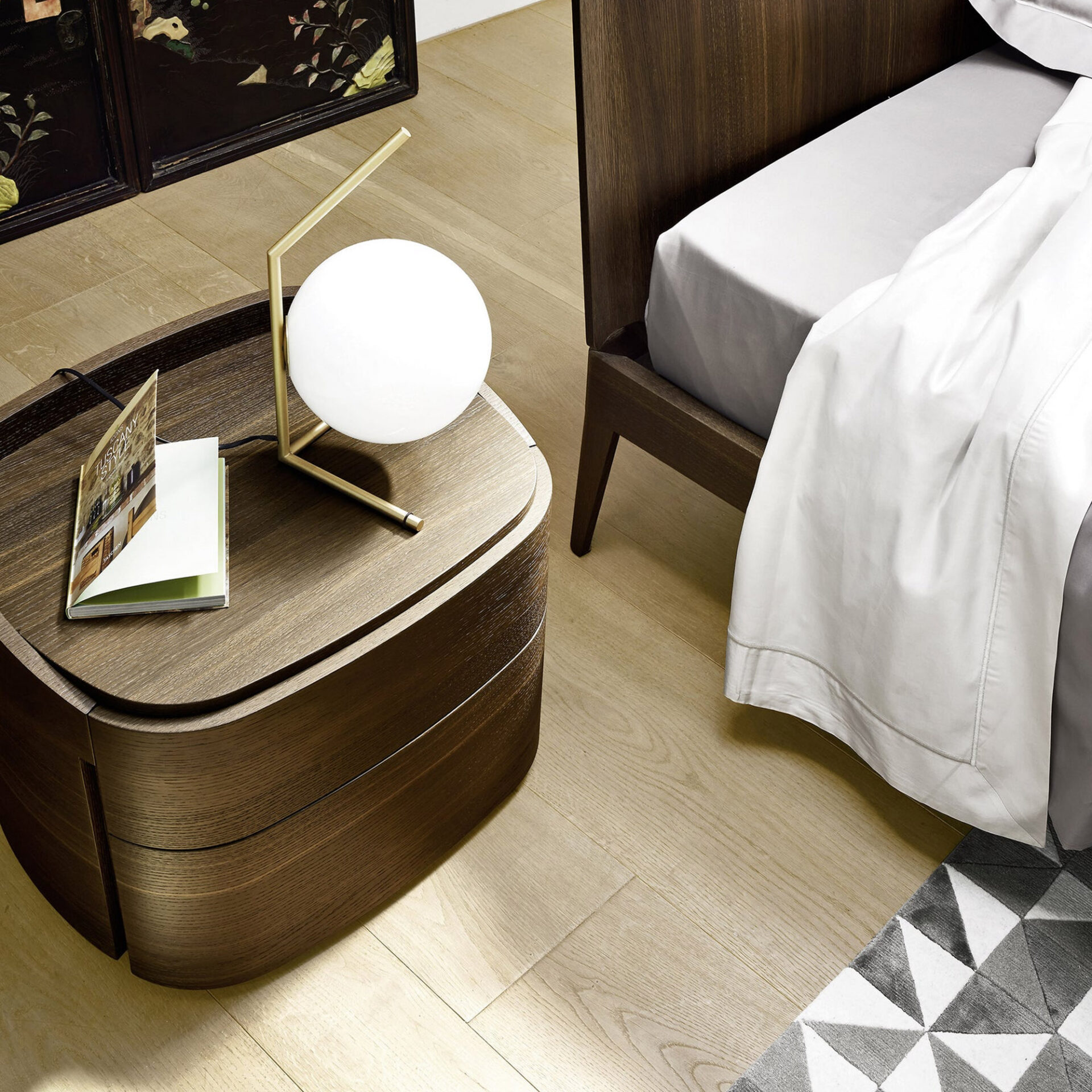 A modern bedroom scene featuring a dark wood curved nightstand with a brass and white spherical lamp and an open book. A bed with light grey and white linens is next to it. The floor is light wood, and a grey and white geometric rug is visible. A decorative dark screen is in the background.