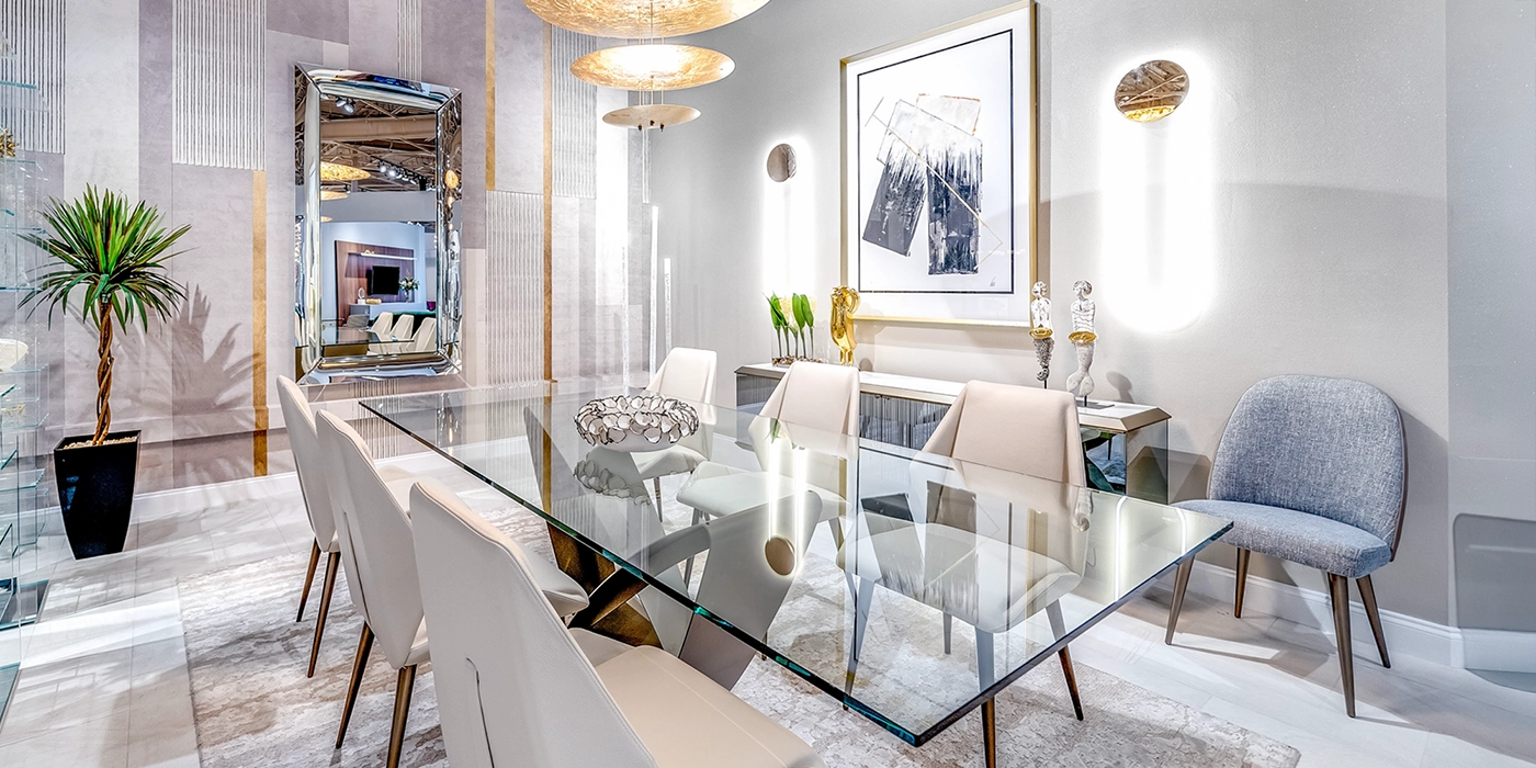A modern and luxurious dining room with a glass table, cream upholstered chairs, a large mirror, abstract art, and gold-toned decorative elements.