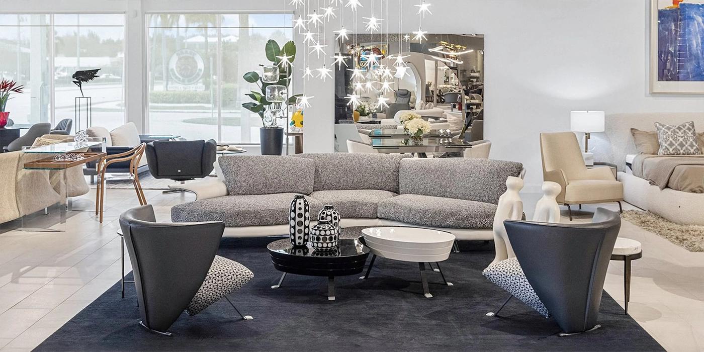 A bright, modern furniture showroom featuring a gray sectional sofa, two coffee tables, and dark gray armchairs on a dark rug, with a starburst chandelier and other contemporary furniture in the background.