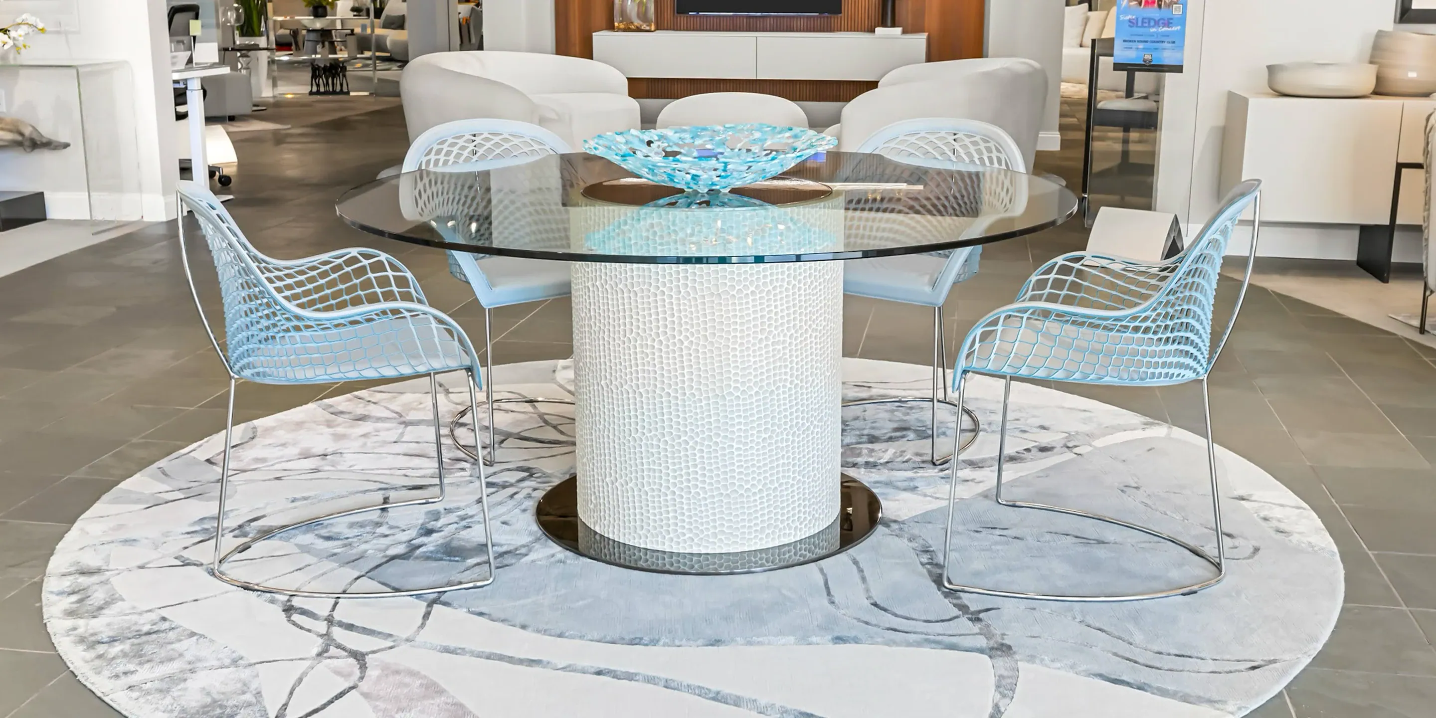 showroom-vignette-oct-24-dining-room-2 Stylish glass dining table with blue woven chairs in contemporary living space.