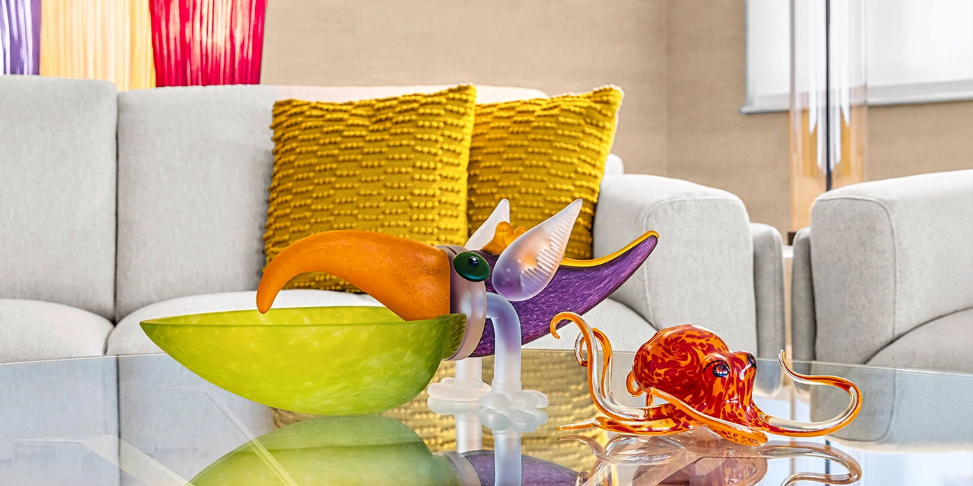 Colorful glass sculptures, including a bird-like figure and an orange octopus, on a glass coffee table in a modern living room with a gray sofa and yellow pillows.