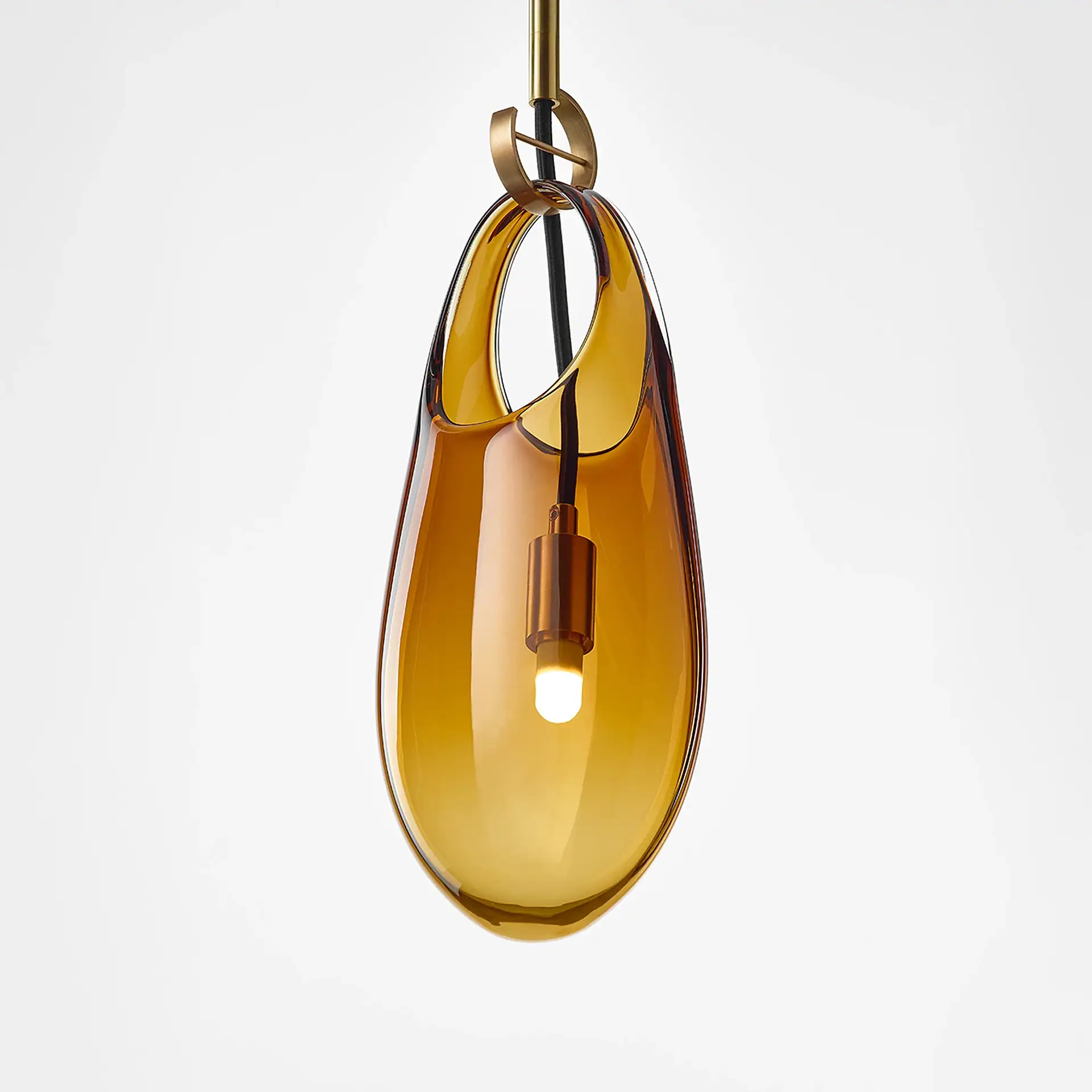 A modern pendant light with an amber-colored, teardrop-shaped glass shade, brass hardware, and a glowing bulb, set against a white background.