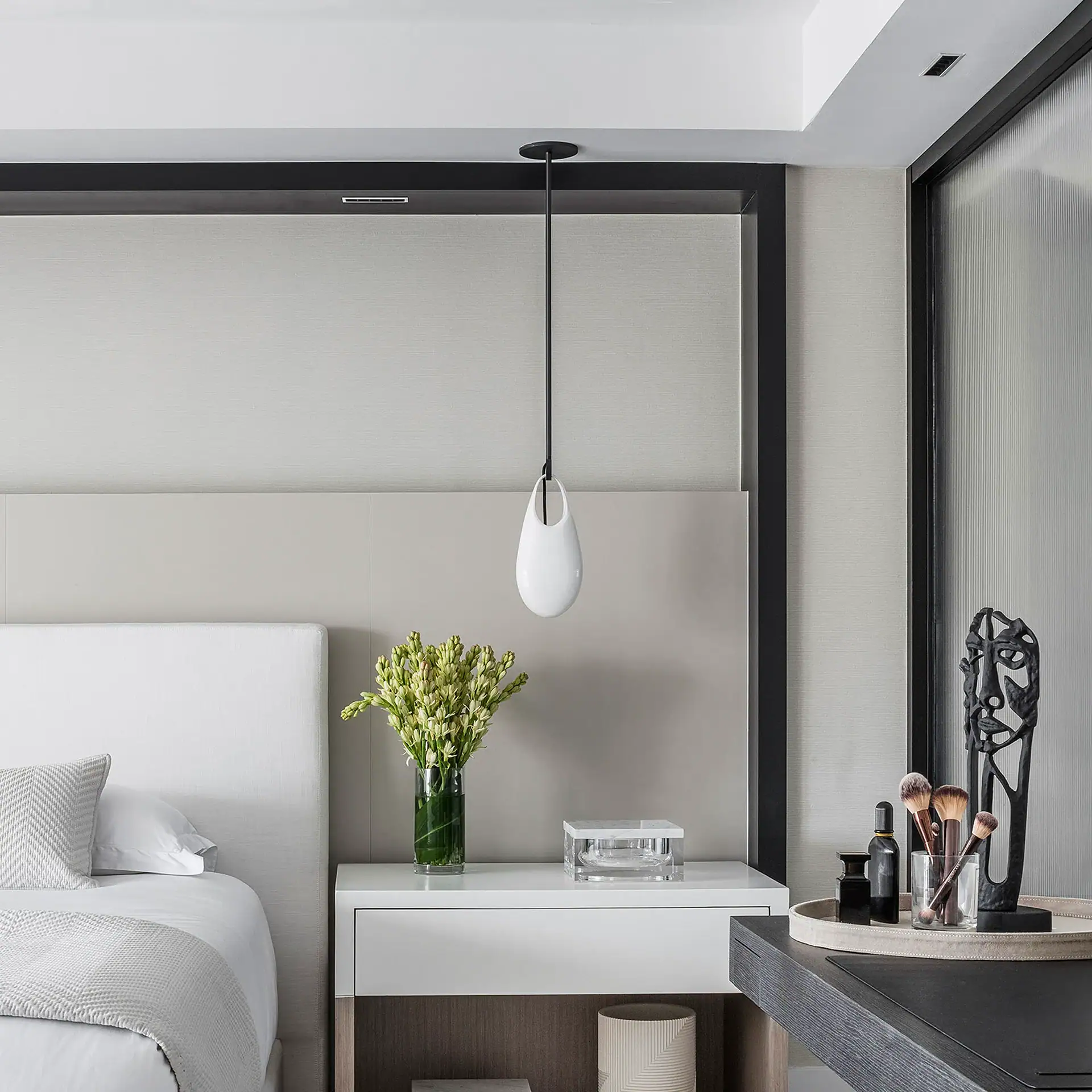 A modern bedroom with a white bed, a white nightstand with flowers, and a dark grey vanity with makeup brushes and an abstract sculpture.