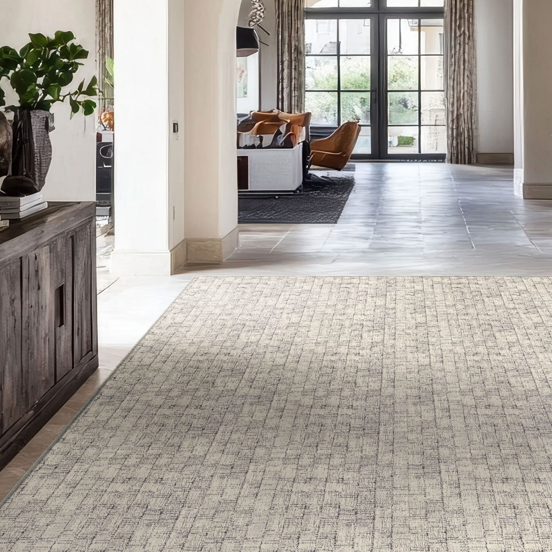 A modern, bright entryway with a light-colored, textured area rug on a stone floor. A dark wood console table with a green plant is on the left, and a bright living room with large, dark-framed glass doors is visible through an archway in the background.