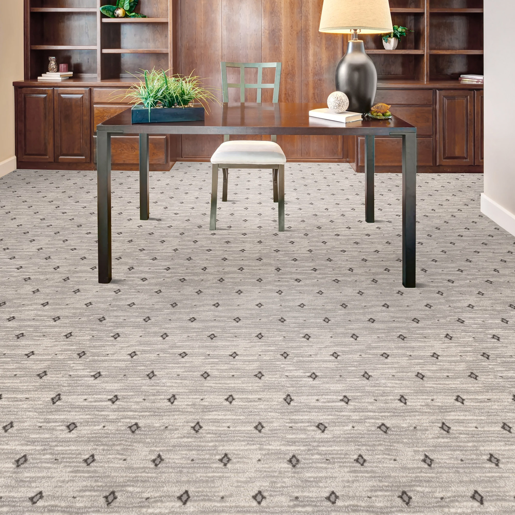 A room with a light gray patterned carpet featuring small dark diamond patterns. A dark wood table with metal legs, a white stool, and decorative items sits in the center. Dark wood cabinetry lines the background.