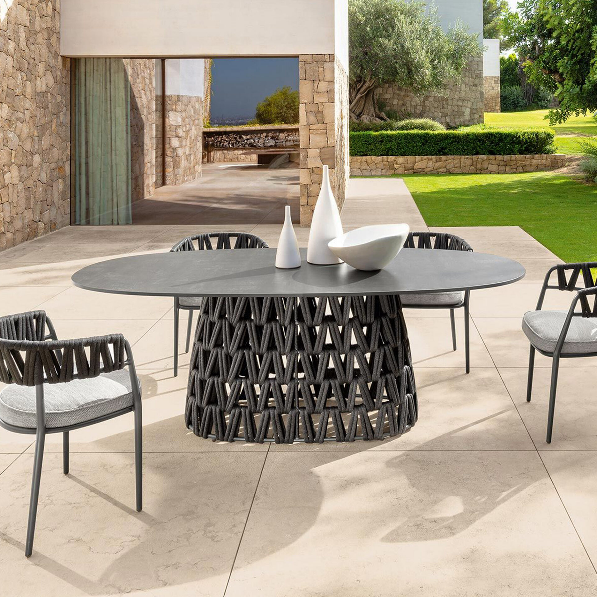 Modern outdoor dining set with an oval dark gray table and woven rope base, surrounded by four matching chairs with light gray cushions, on a tiled patio next to a stone building and lush green garden.