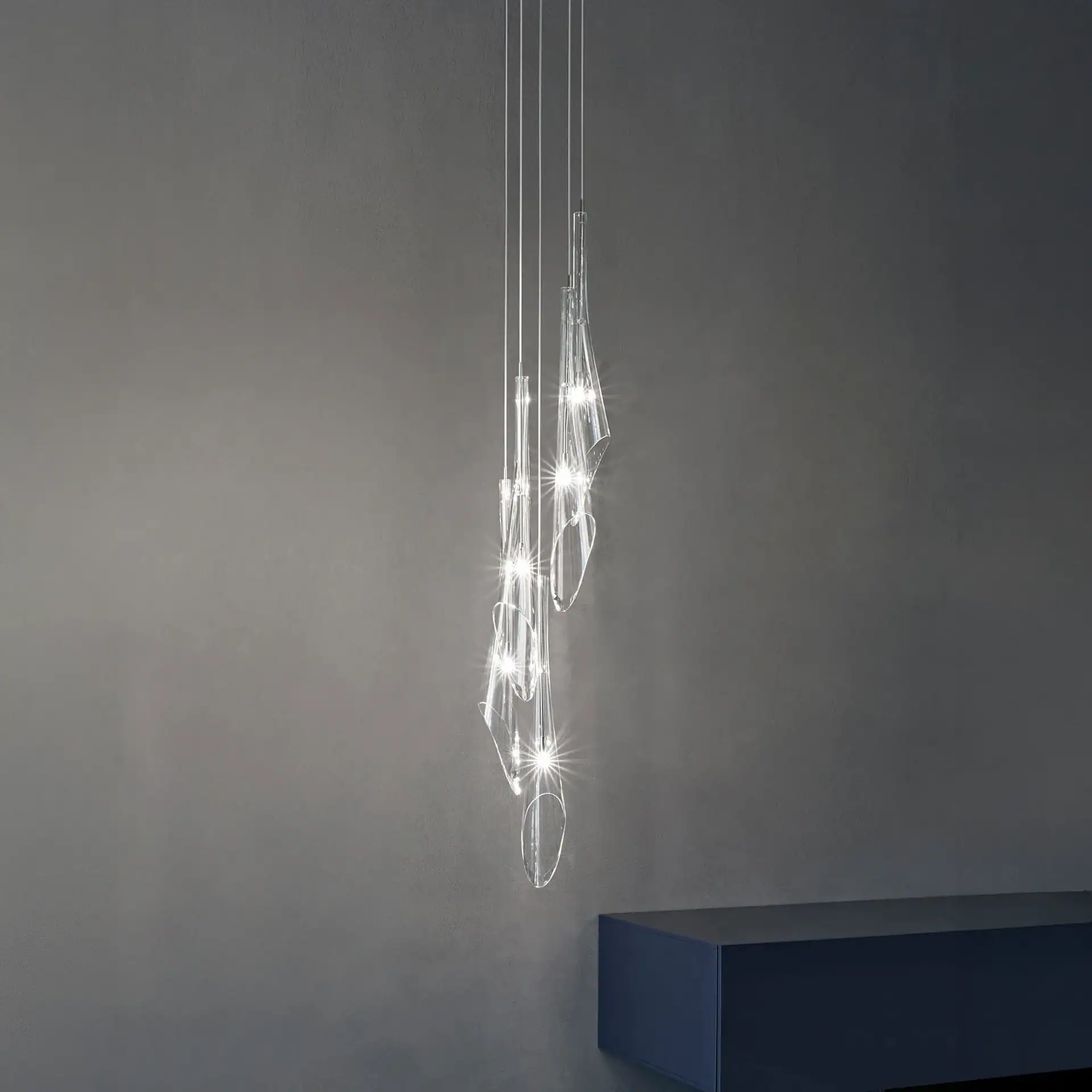 A modern pendant light fixture with multiple clear, illuminated elements hanging at different lengths against a textured grey wall, with a dark blue floating cabinet visible at the bottom right.