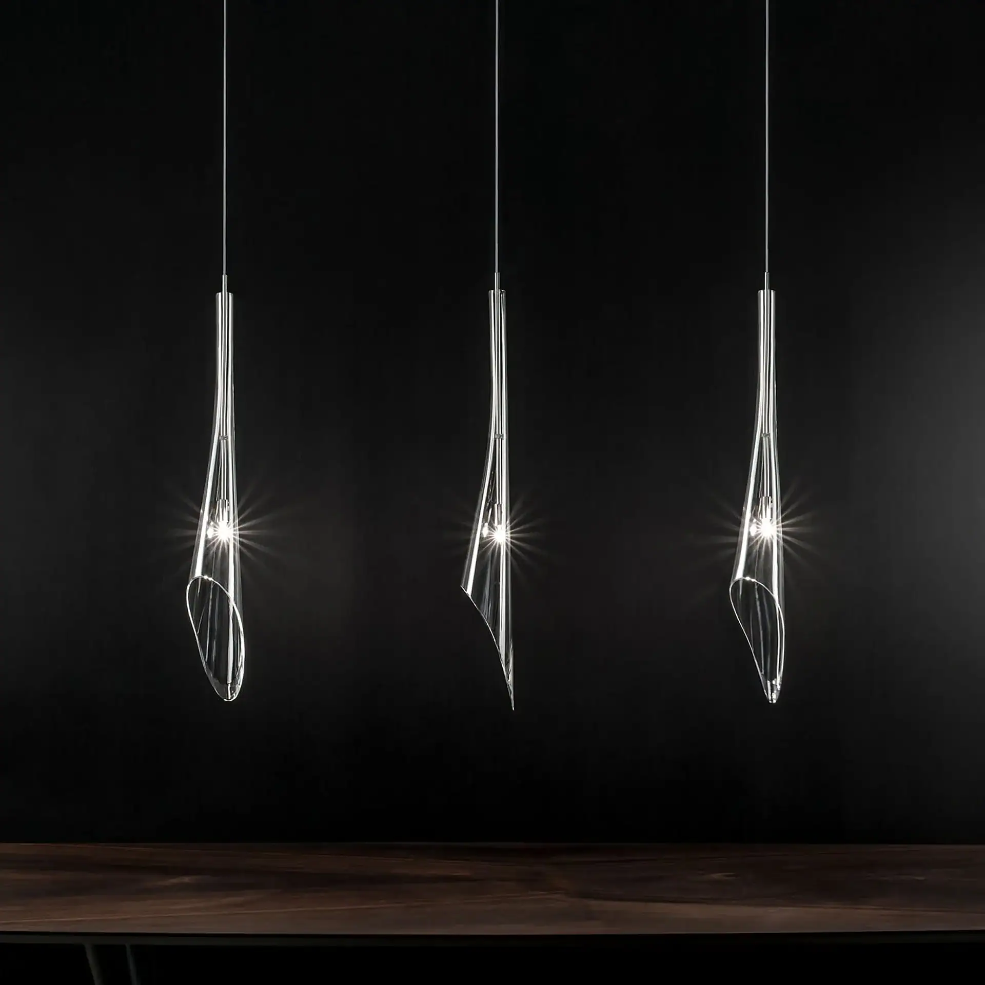 Three illuminated, clear, twisted glass pendant lights hang against a dark background, with a wooden surface below.