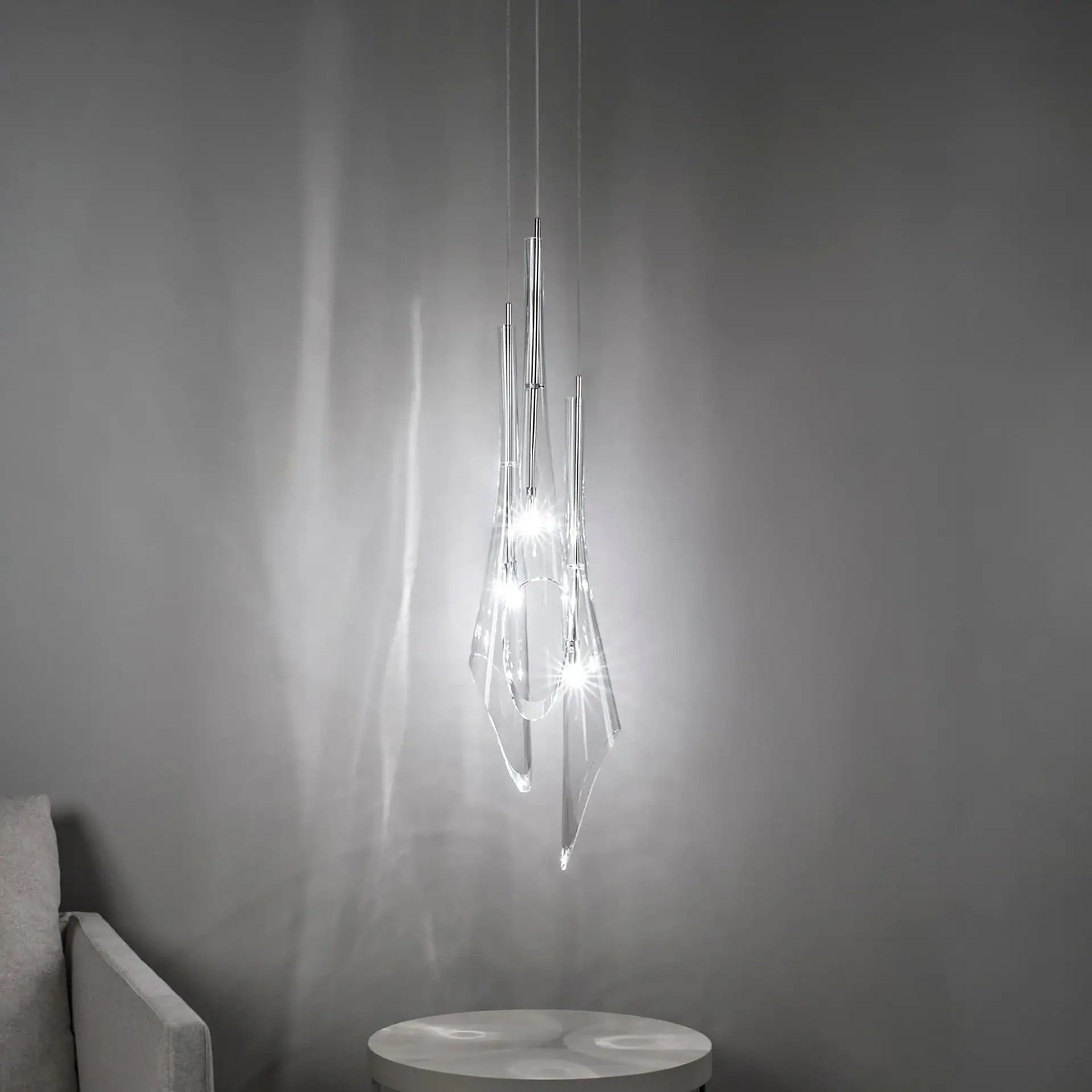 A modern, clear sculptural pendant light fixture hangs in a minimalist room, casting light patterns on a gray wall and a round table.