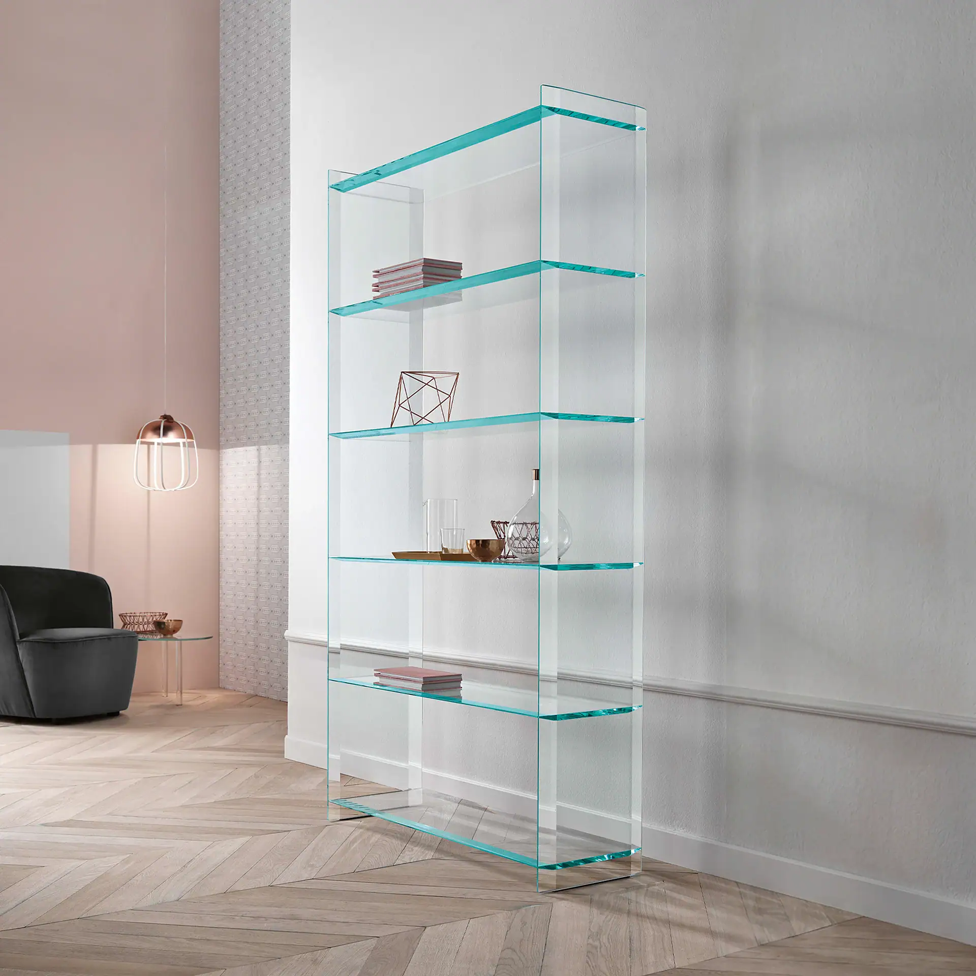 A tall, clear glass bookshelf with five shelves, displaying books and decorative items, in a modern living room with pink and white walls, a grey armchair, and a herringbone wood floor.
