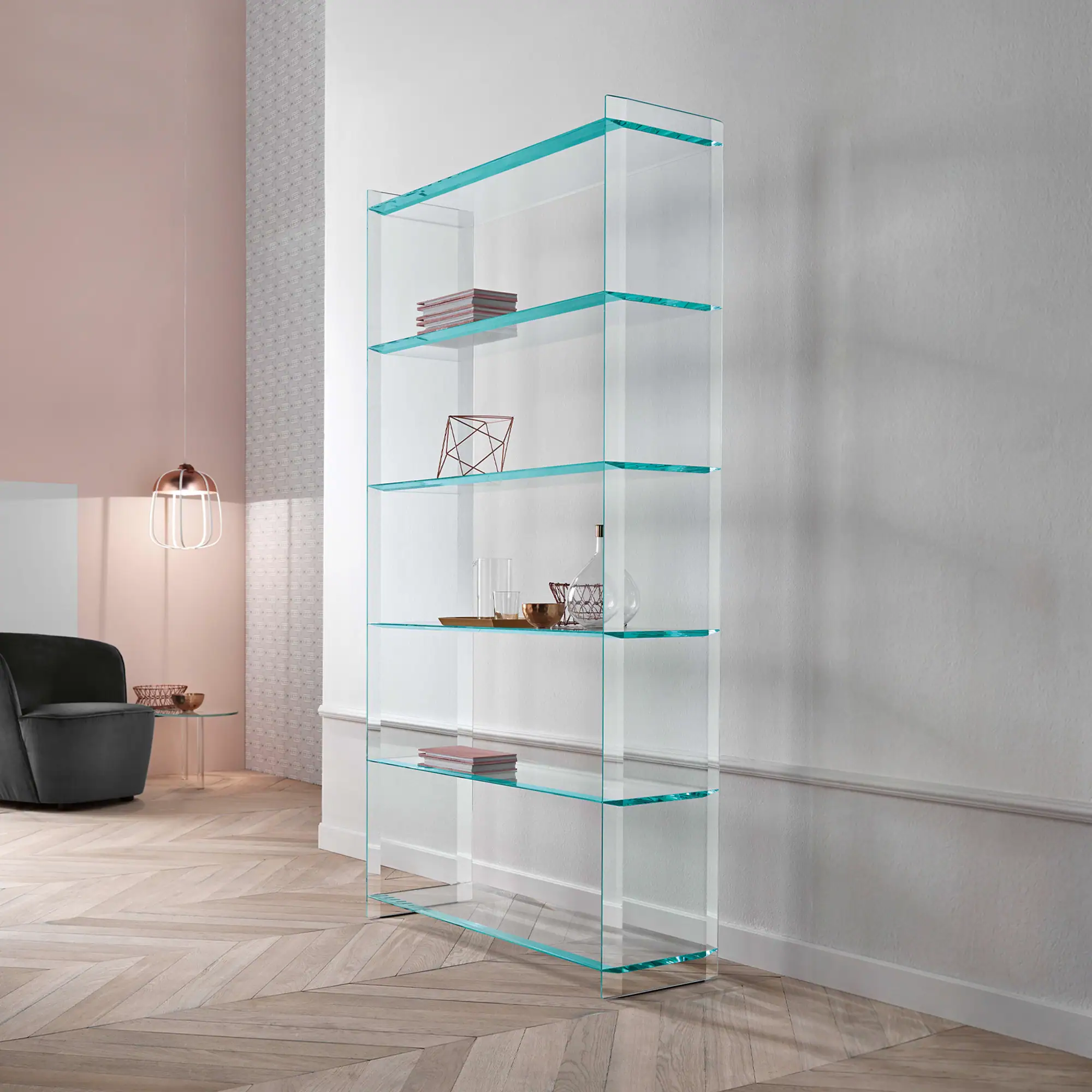 A tall, clear glass bookshelf with five shelves, displaying books and decorative items, in a modern living room with pink and white walls, a grey armchair, and a herringbone wood floor.