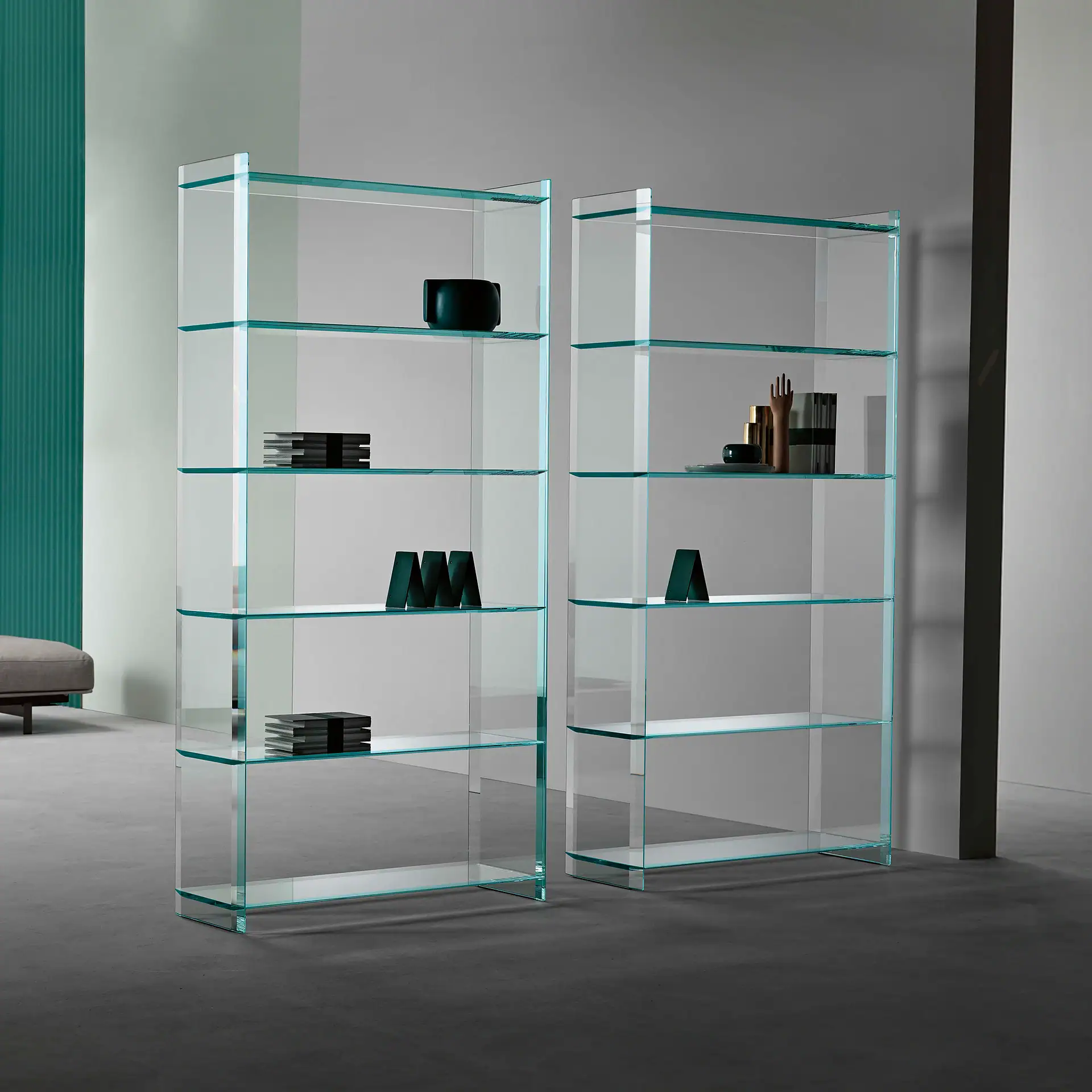 Two tall, clear glass bookshelves with a blue-green tint, displaying various decorative objects in a minimalist room.